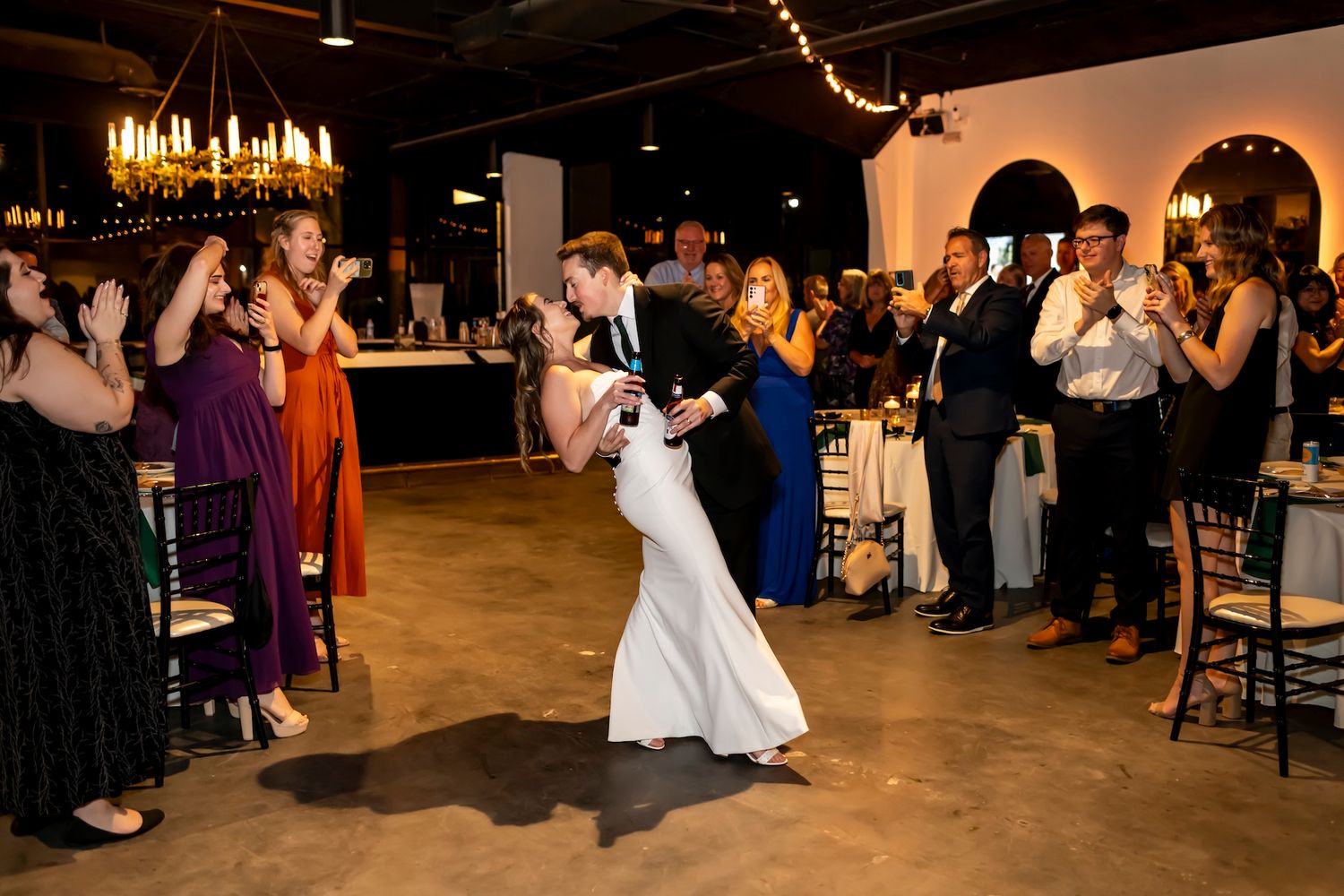 Wedding guests watch from sidelines as newlyweds dance in an elegant reception venue with chandelier lighting overhead.
