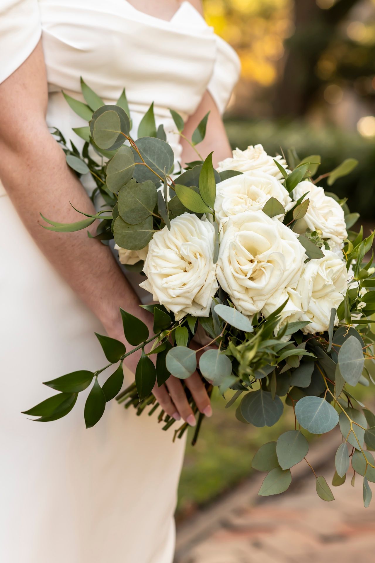 Close-up of a bridal bouquet featuring white roses and eucalyptus leaves against a white dress.