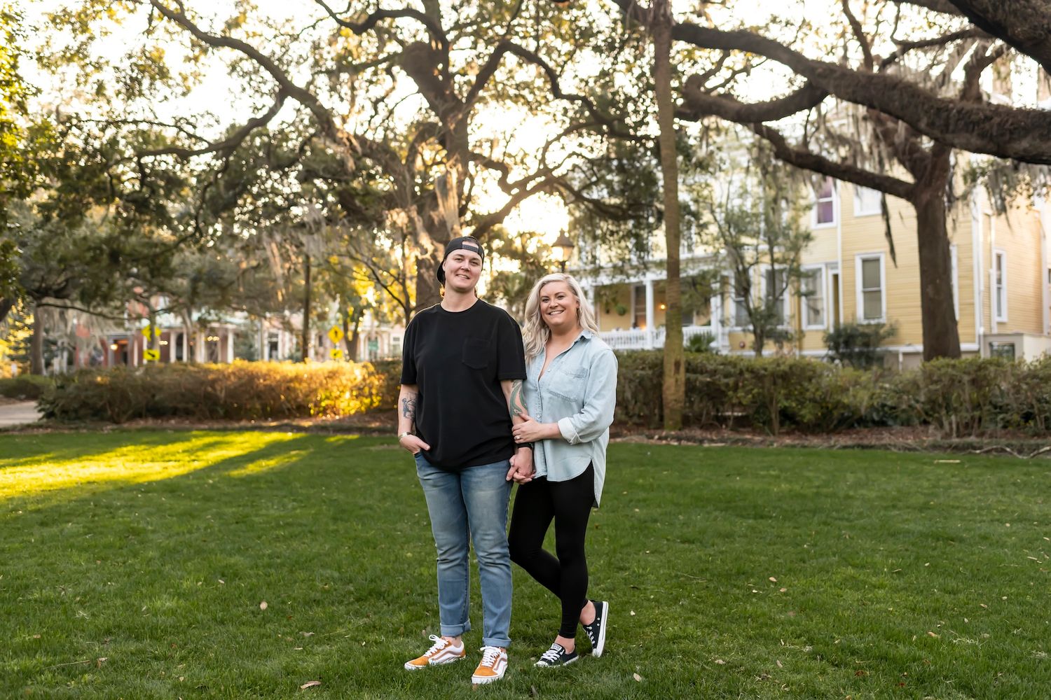 Two people stand together on a grassy lawn during golden hour with historic buildings and Spanish moss trees in background.