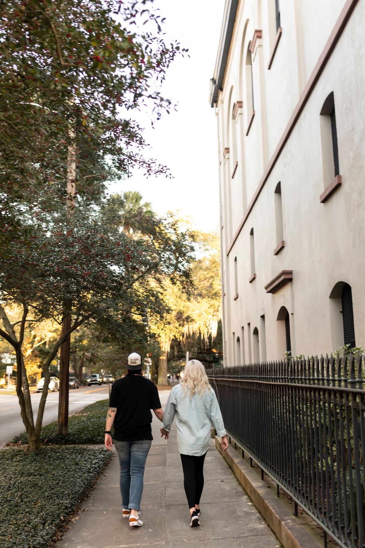 A couple walks hand in hand down a historic sidewalk with white building and iron fence in Charleston.