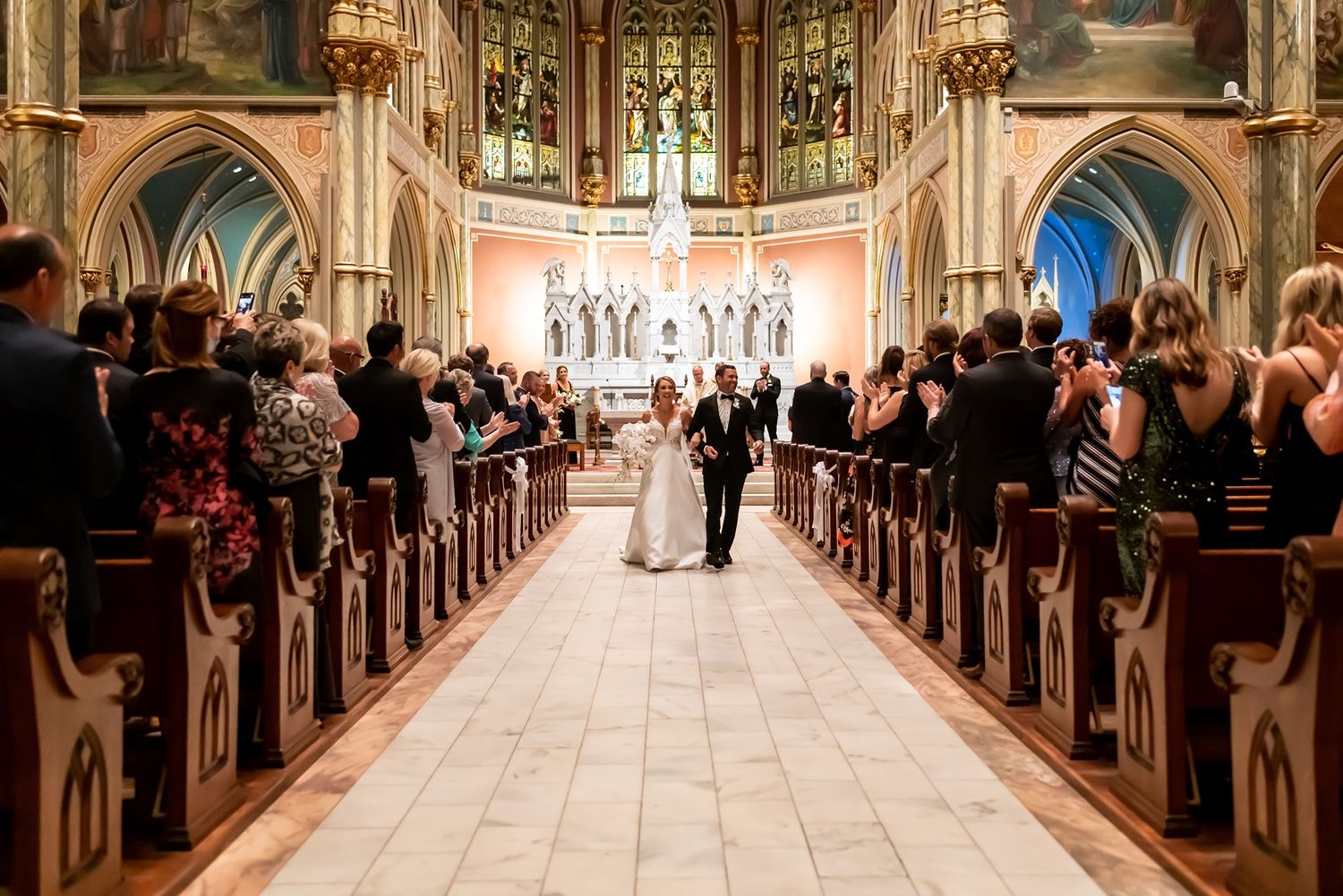 A couple stands at the altar during their wedding ceremony in an ornate Gothic cathedral with stained glass windows.