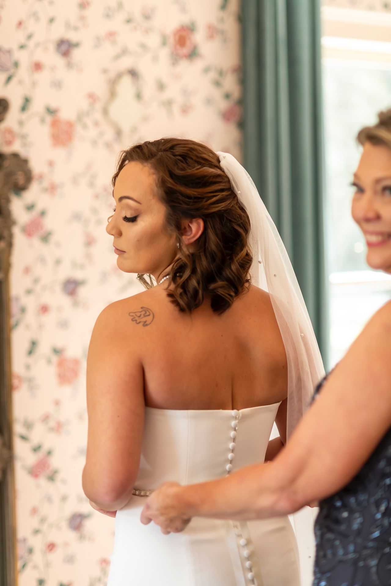 A woman in a white wedding dress with buttons down the back stands as someone helps with final adjustments.