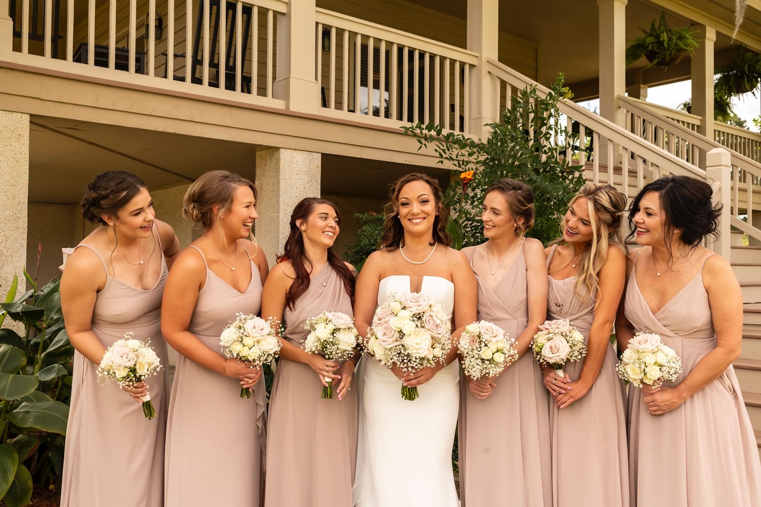 Seven women in matching blush pink bridesmaid dresses and white bouquets pose with bride in white on porch.