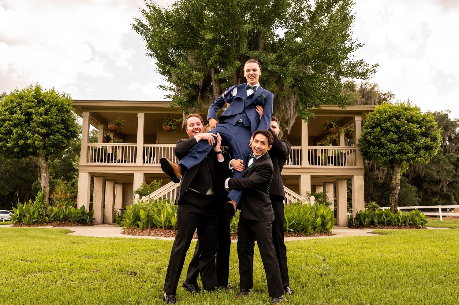 Group in matching dark suits posing together on green lawn in front of two-story residential building with porch.