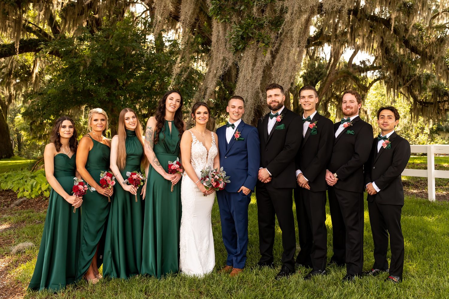 Wedding party in formal attire poses outdoors with bridesmaids in green dresses and groomsmen in black suits.