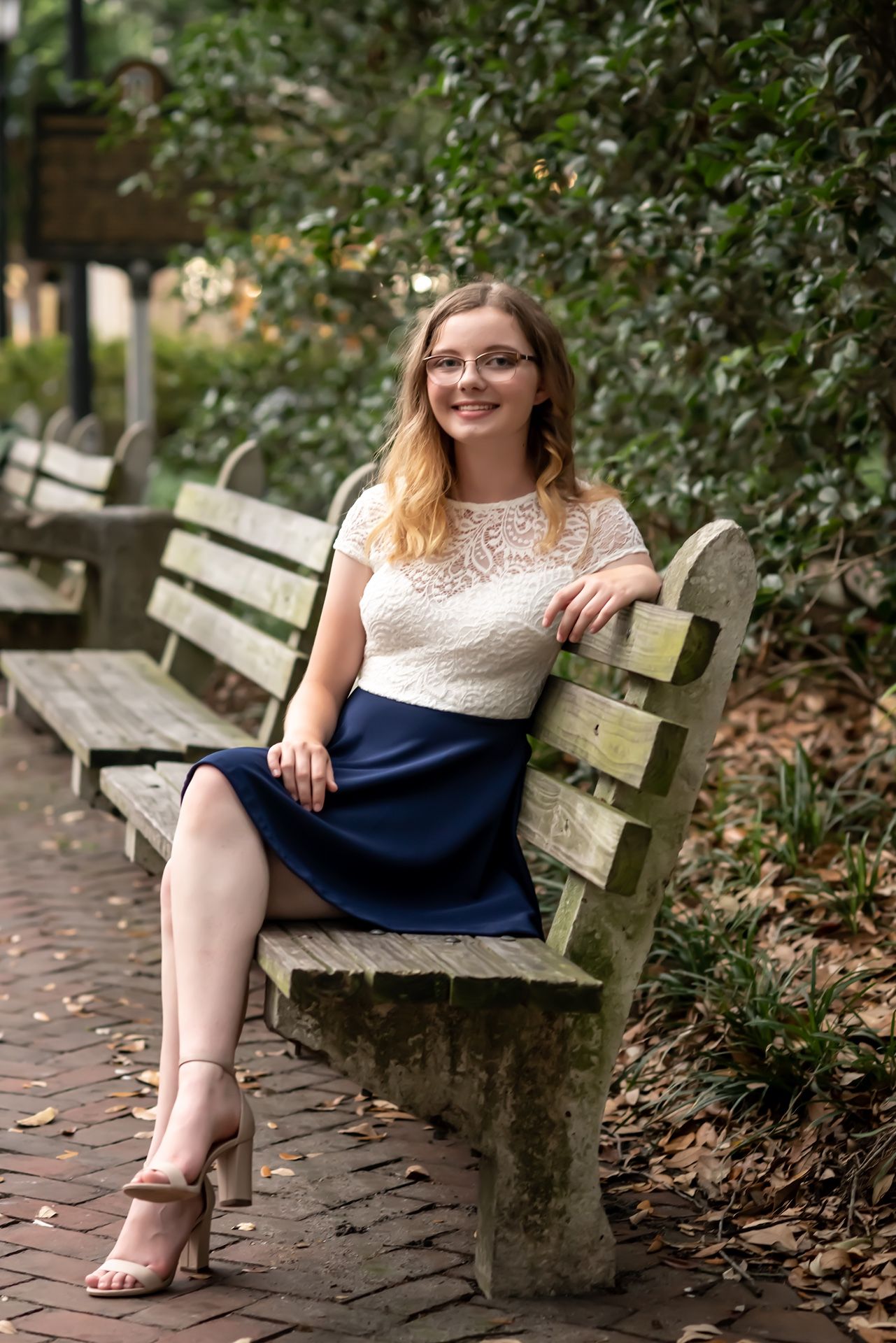 A woman in a white lace top and navy dress sits at different angles on a rustic wooden garden bench surrounded by foliage.