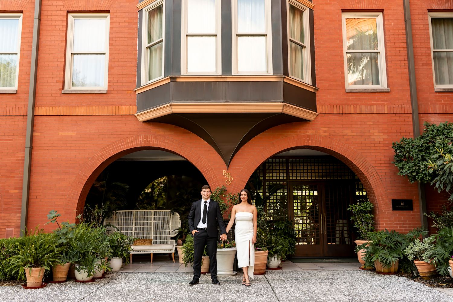 A couple stands in front of a rustic brick townhouse with an arched entrance and bay window in an urban setting.
