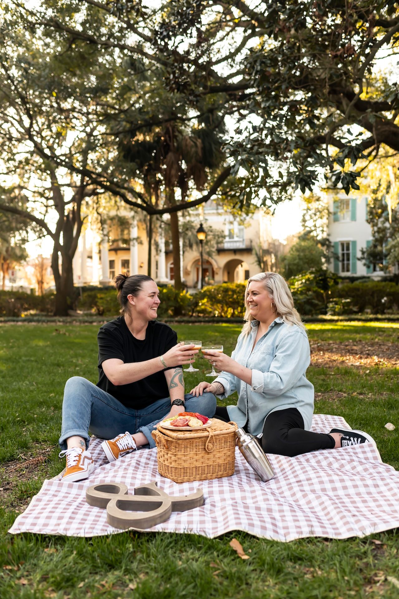 Two people enjoying a picnic on a checkered blanket in a sunlit park with a wicker basket and snacks.