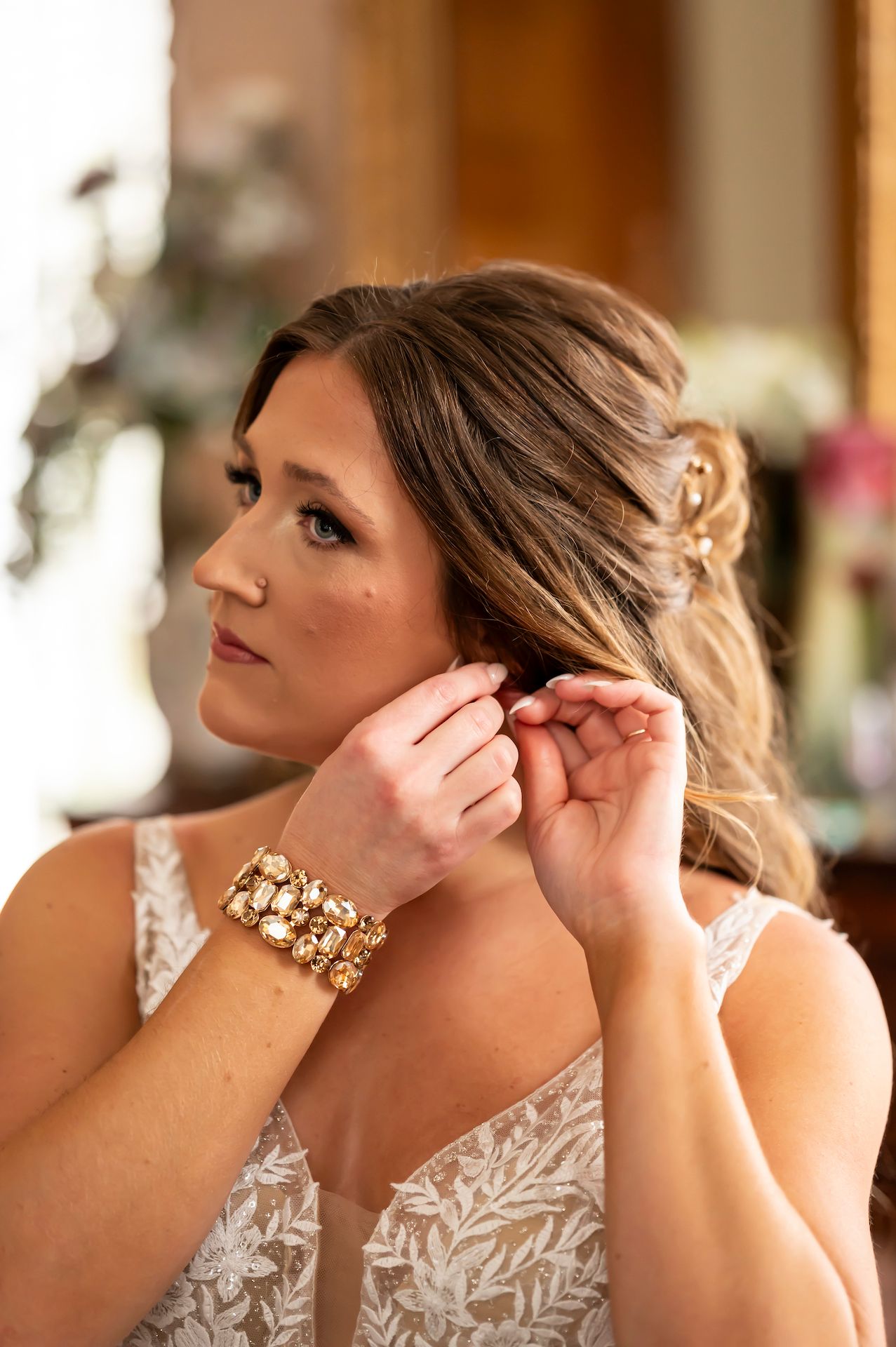 Bride puts on earrings while wearing a lace wedding dress and gold bracelet in warm natural lighting.