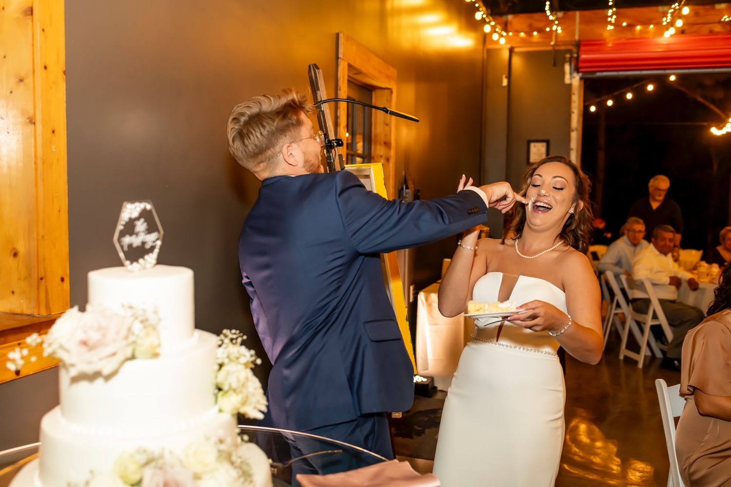 Newlyweds share a playful moment while cutting their wedding cake at an intimate indoor reception with warm lighting.