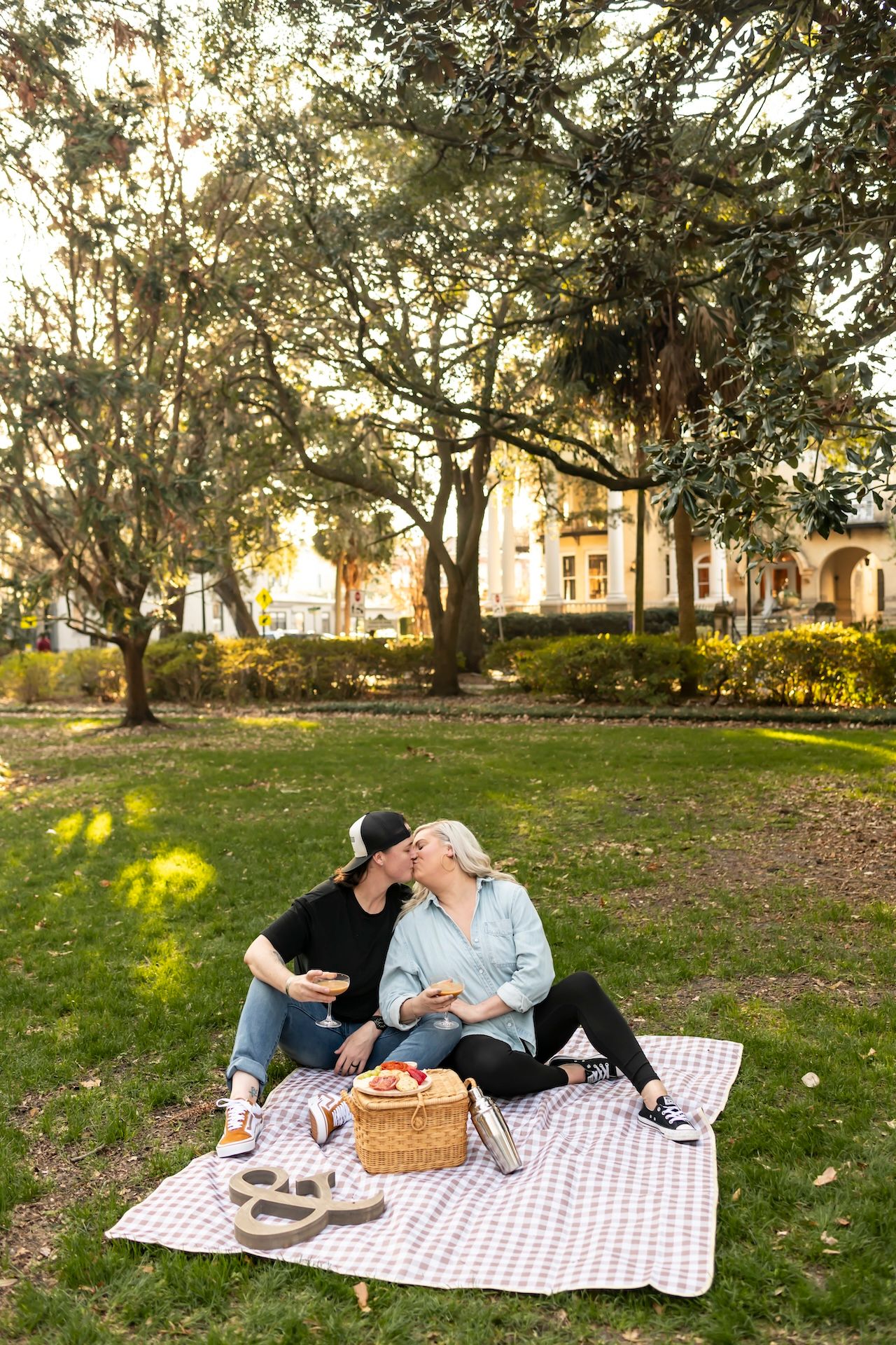 Couple enjoying a romantic sunset picnic on a polka dot blanket with a wicker basket in a grassy park setting.