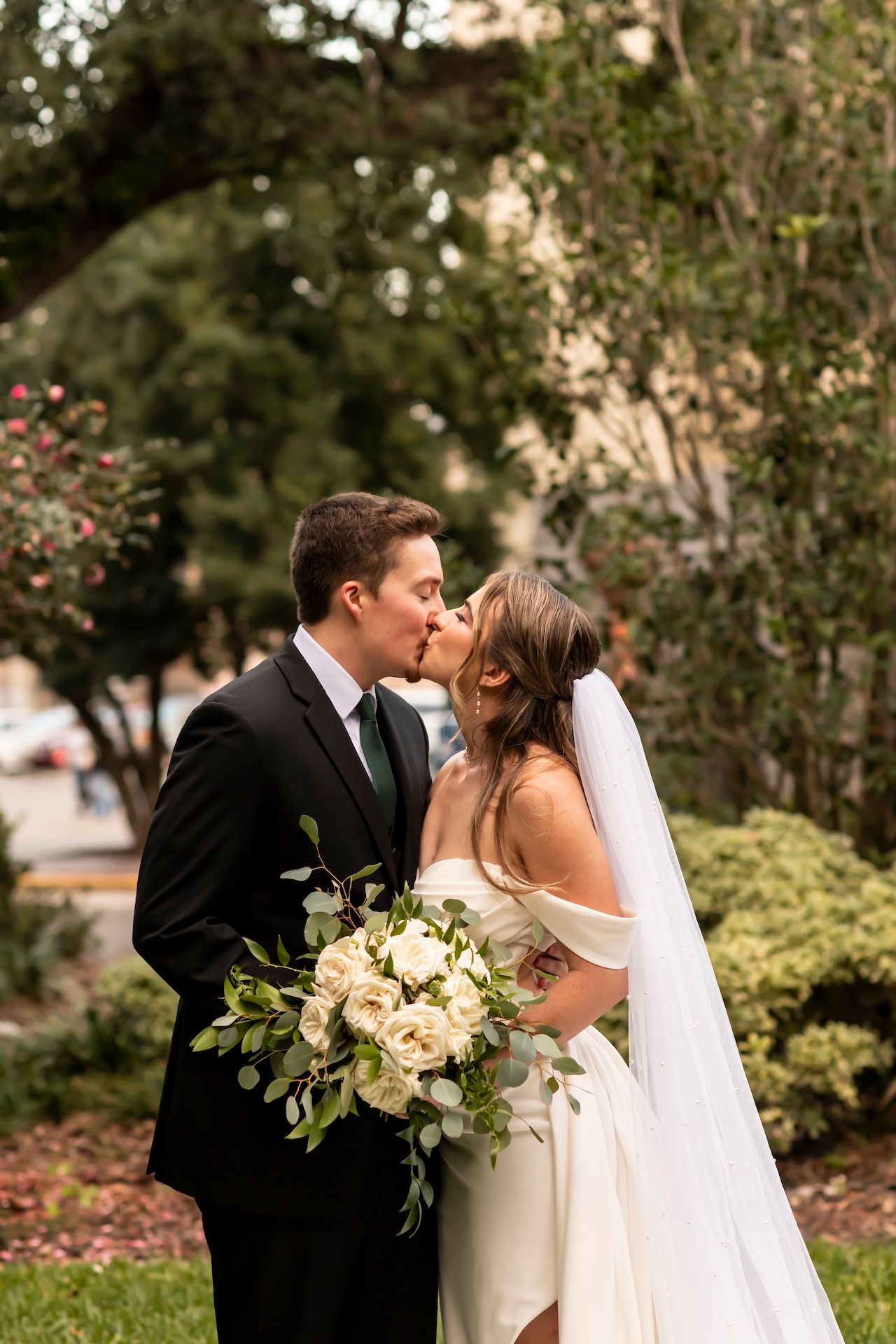 A romantic wedding couple embraces outdoors while the bride holds a white and green floral bouquet.