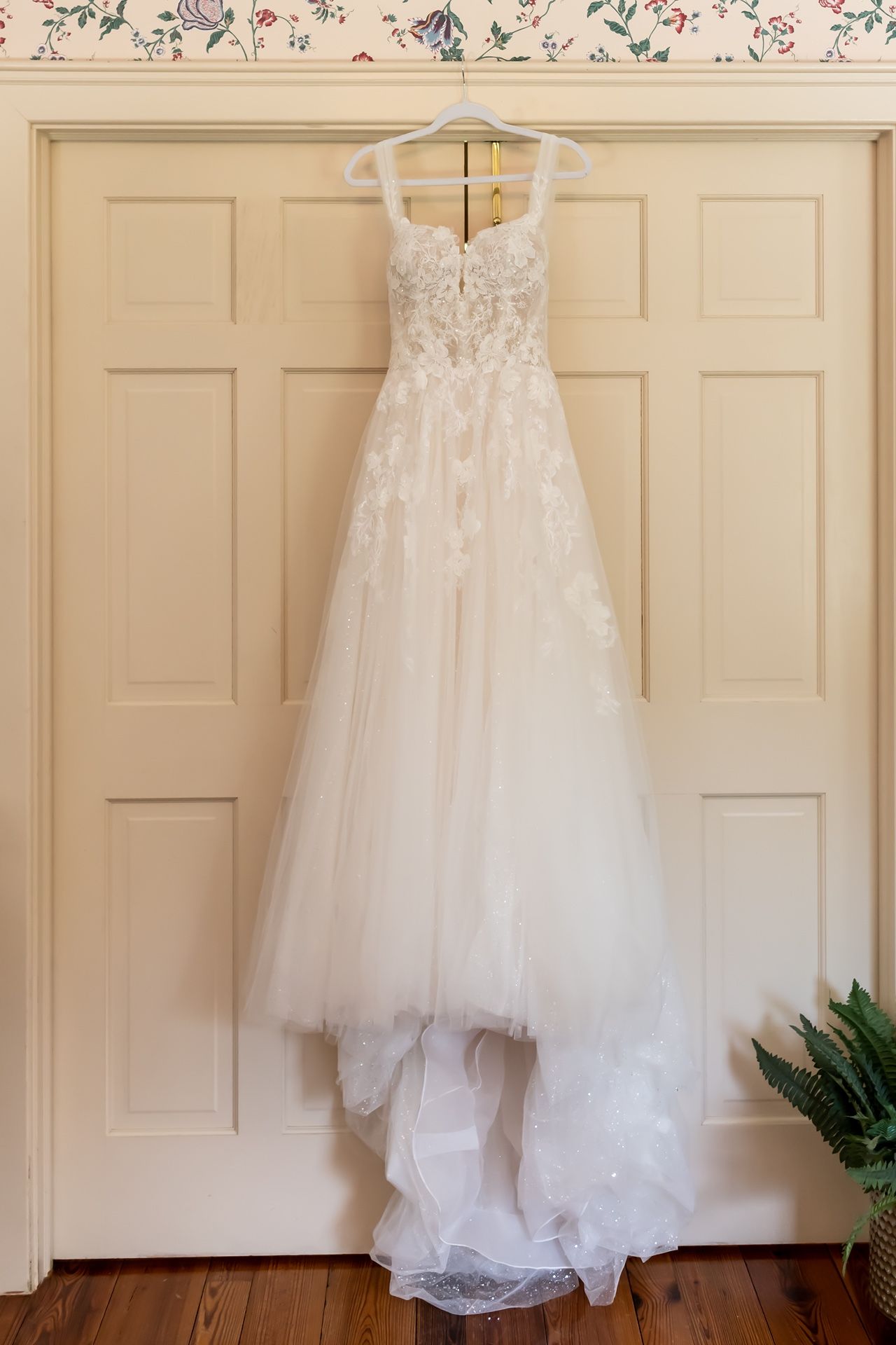 An elegant white lace and tulle wedding gown with sweetheart neckline hangs on a white hanger against a paneled door.