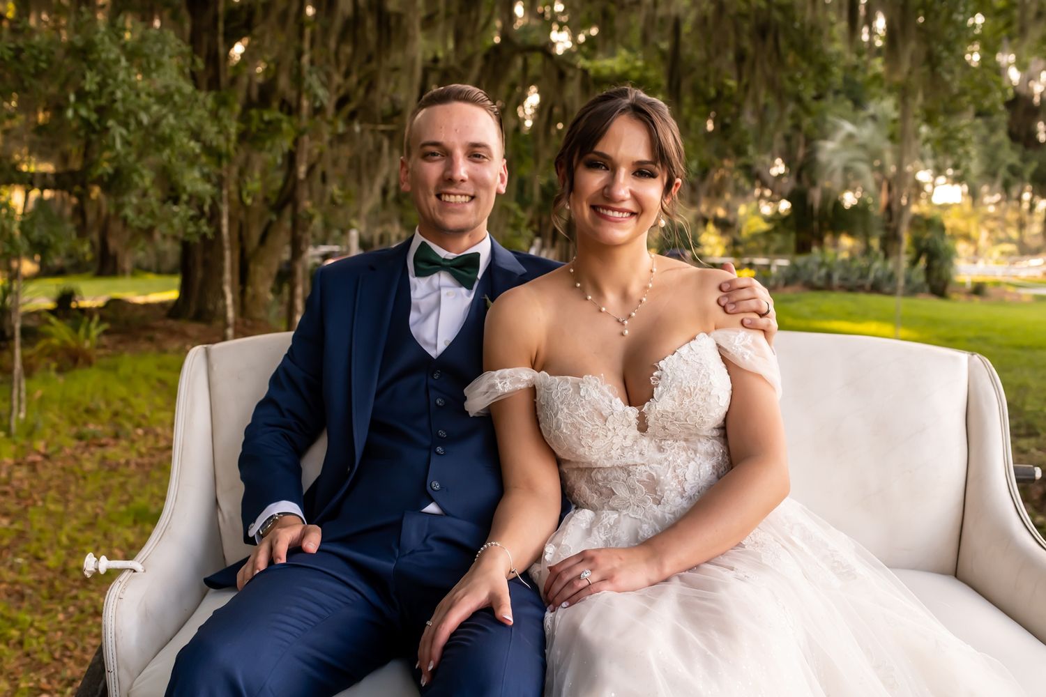 A newlywed couple sits together on an elegant white couch outdoors in a garden setting.