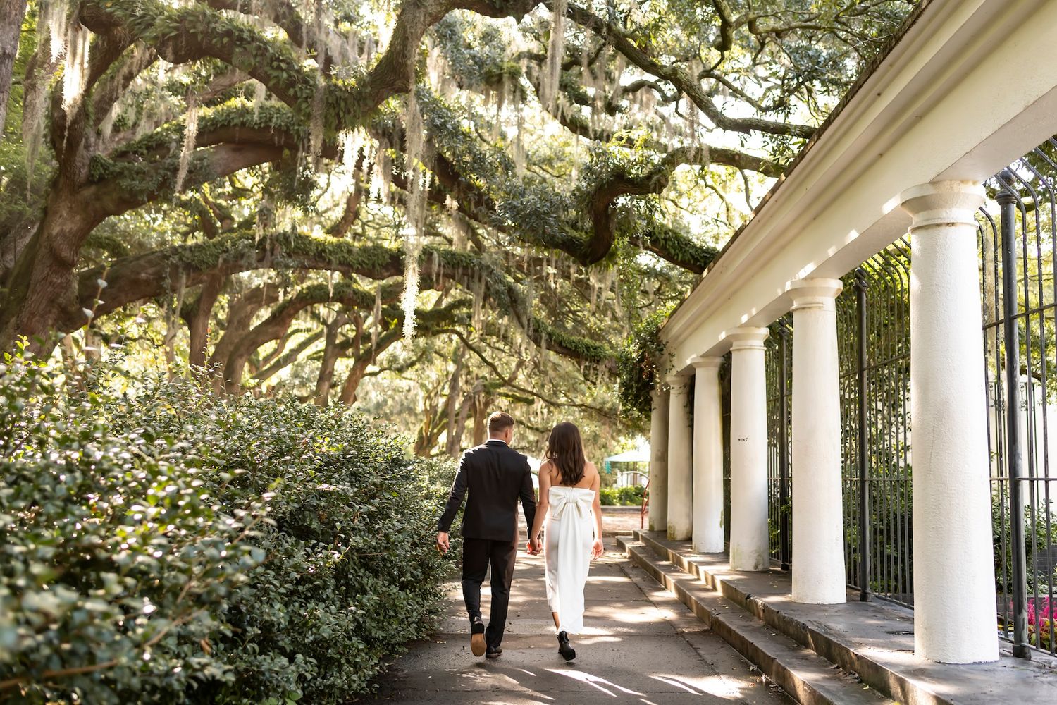 A couple walks down a romantic garden path covered by Spanish moss and lined with elegant white columns.