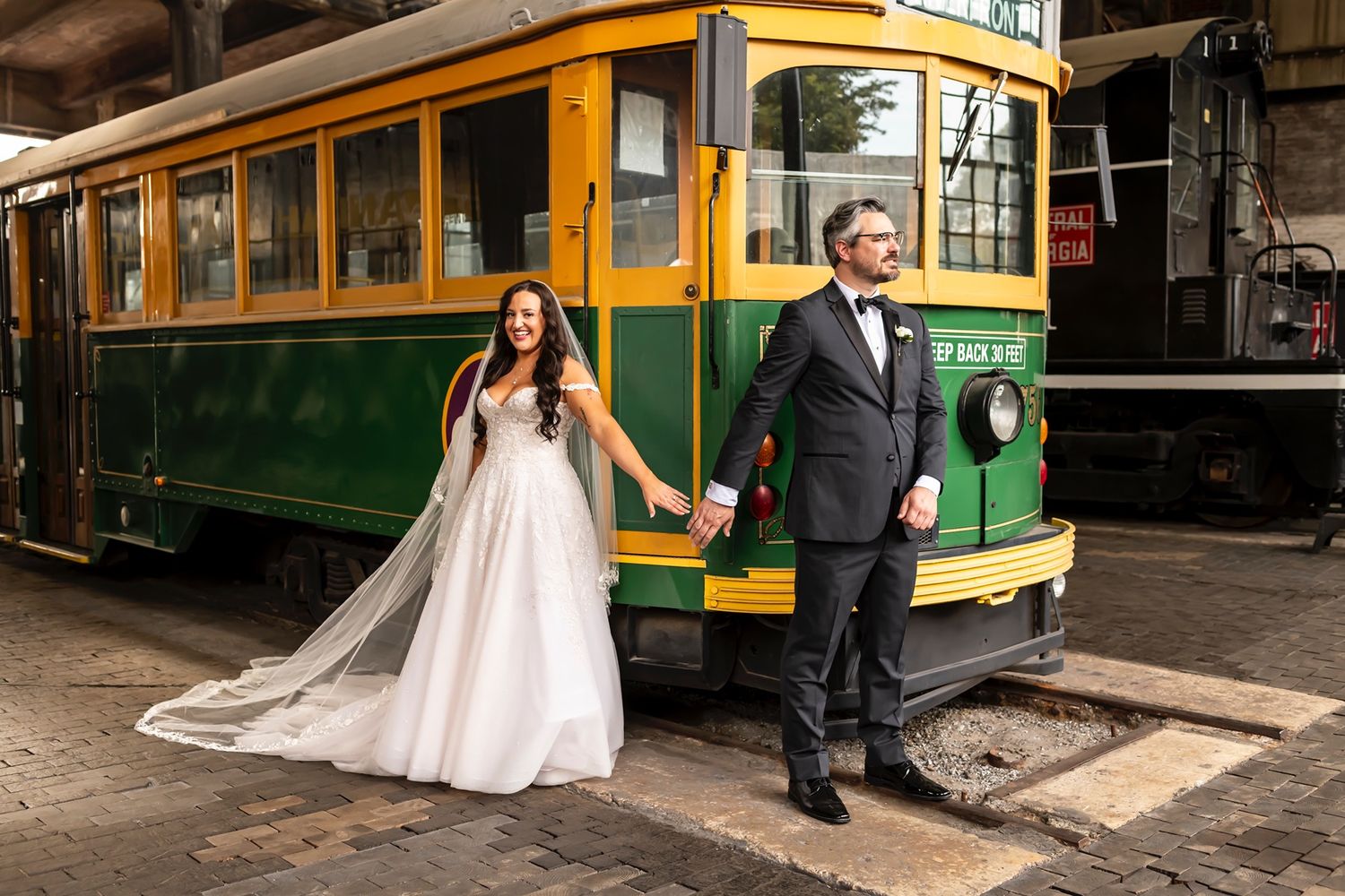 A couple stands hand in hand in wedding attire next to a vintage yellow and green trolley.