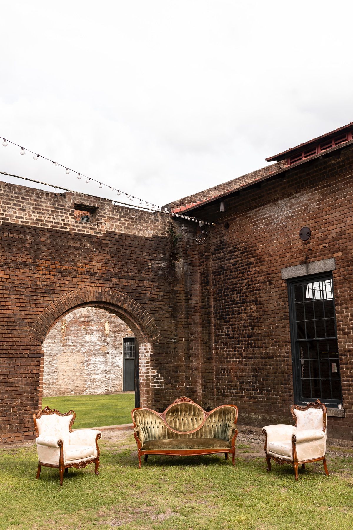 Vintage brick courtyard with antique furniture seating area featuring two upholstered chairs and a wicker settee on grass.