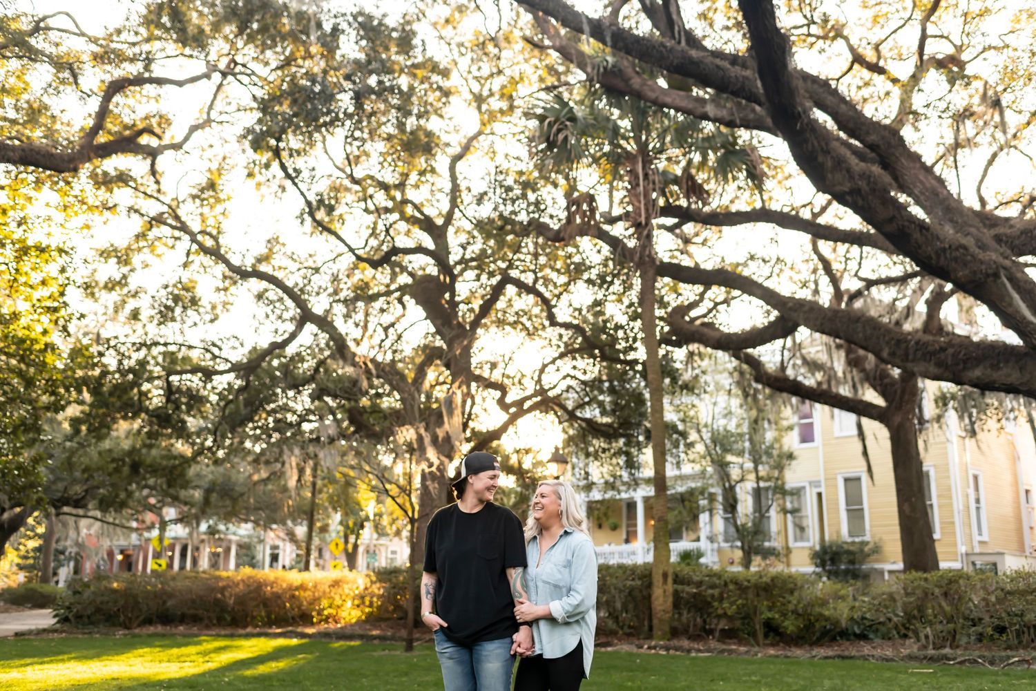 A couple stands beneath sprawling oak tree branches at sunset in a garden setting with historic buildings in background.