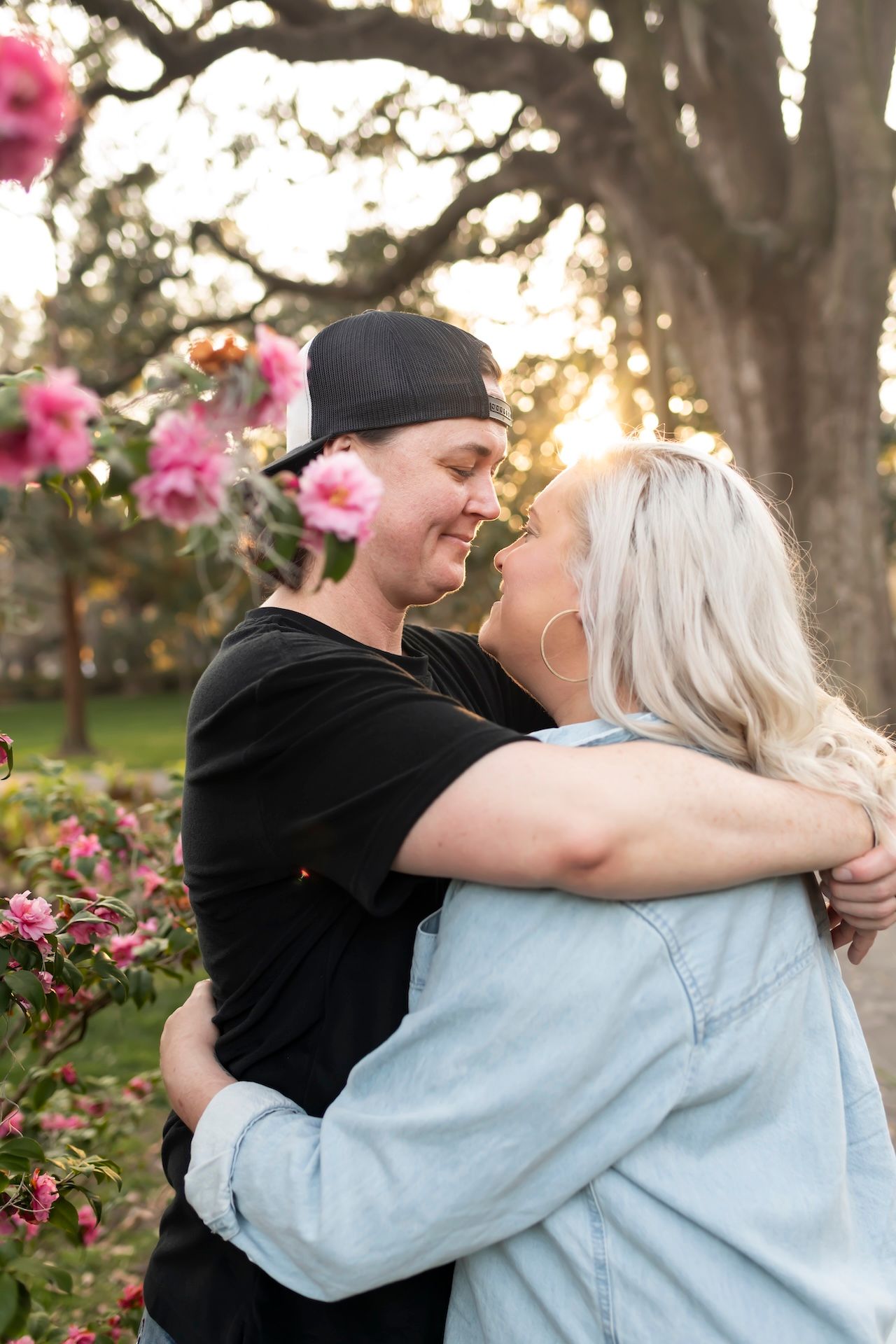 Happy couple embraces in garden with blooming pink roses and sunlit trees in background at sunset.