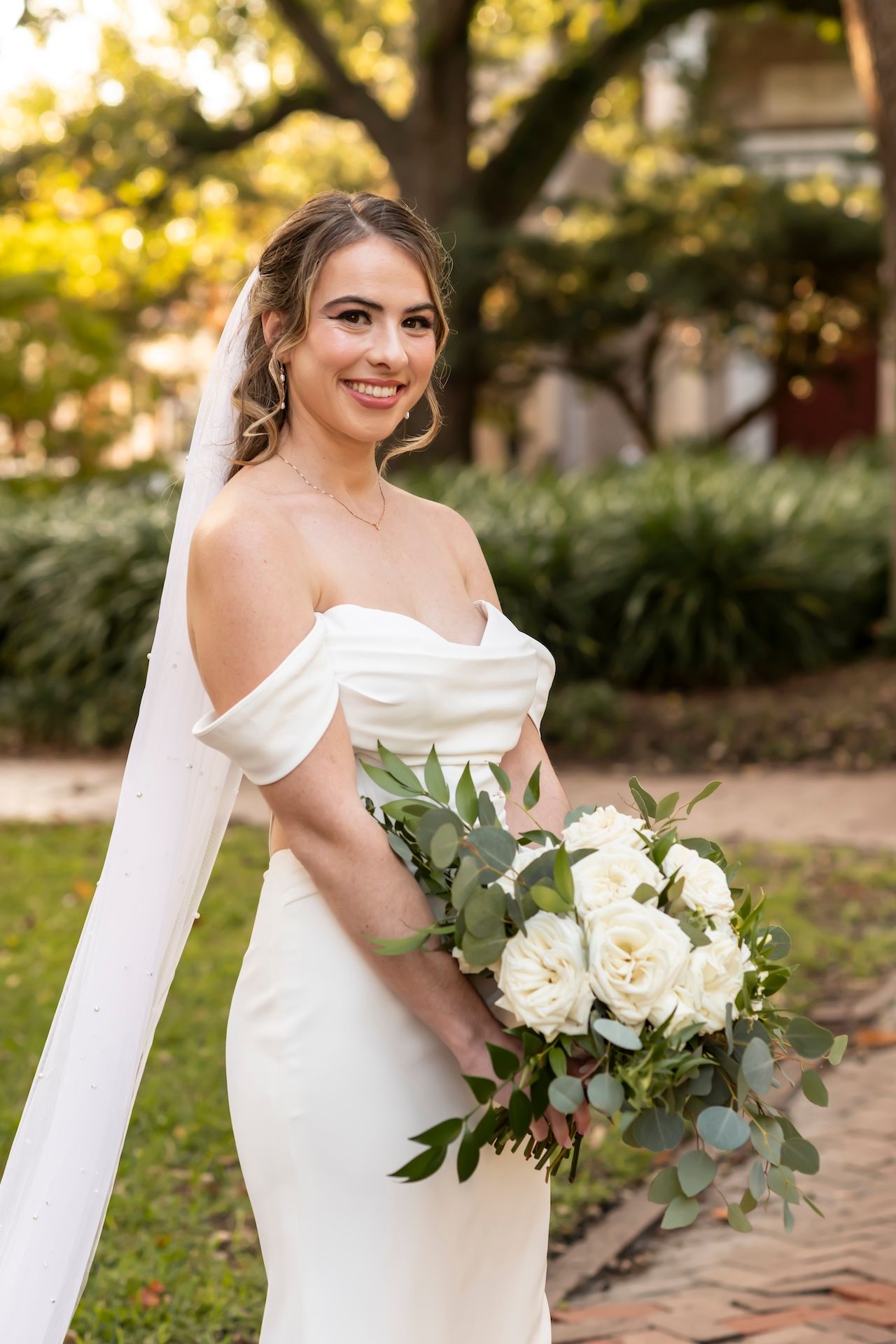 Bride in elegant off-shoulder white dress holds bouquet of white roses and eucalyptus in garden setting.