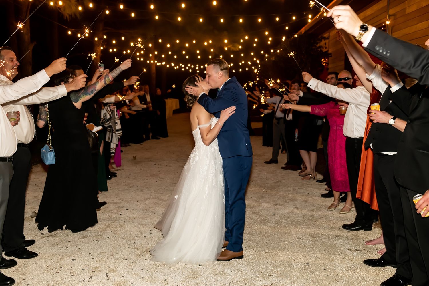 Couple shares romantic dance moment under string lights at night while wedding guests create a walkway with sparklers.