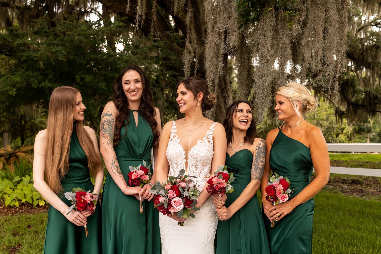 A bridal party stands together outdoors wearing emerald green dresses and holding colorful floral bouquets.