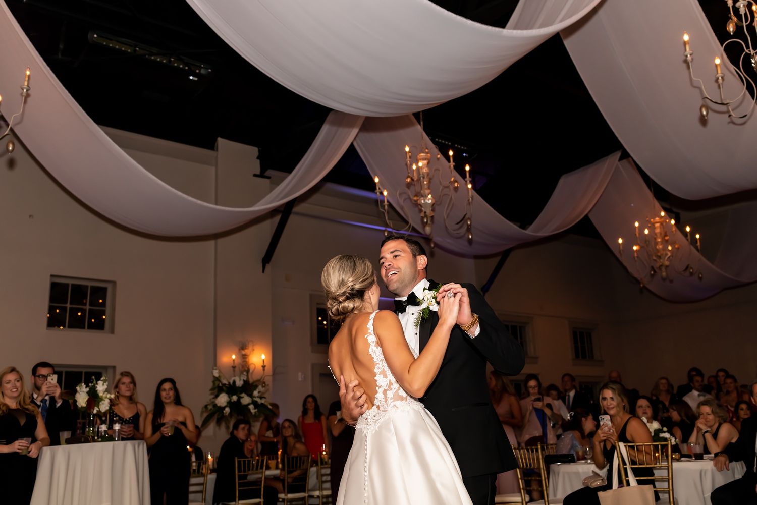 A couple shares their first dance under elegant white draping and twinkling lights at an indoor wedding reception.