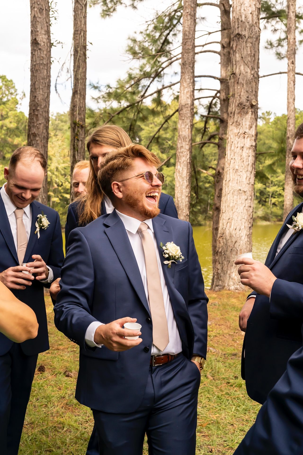 Wedding party members in navy suits mingle and socialize in a wooded outdoor setting during a celebration.