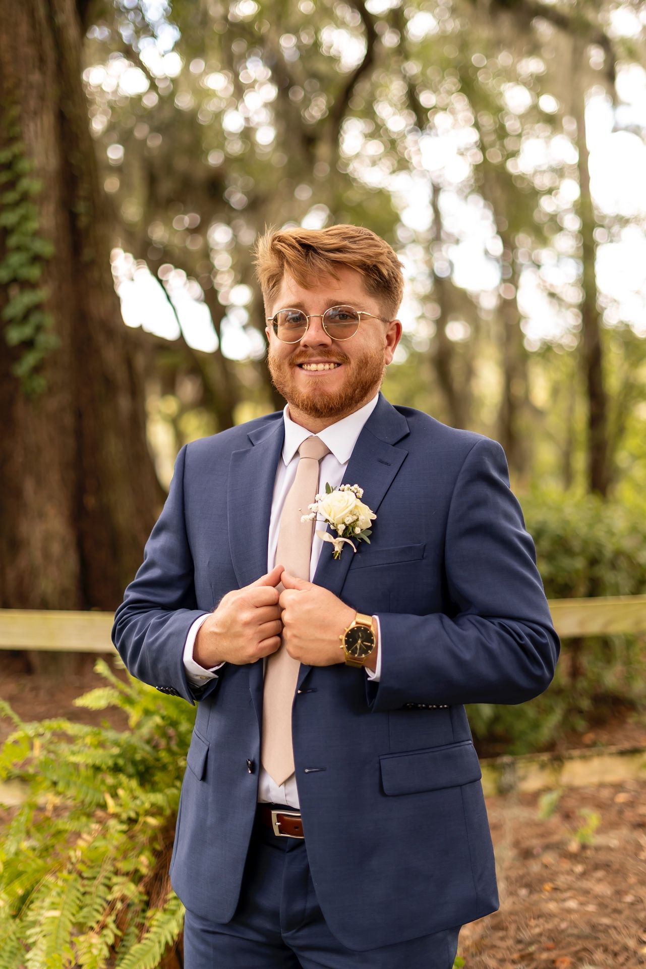 A groom in a navy suit and glasses smiles while adjusting his jacket in a sunlit outdoor setting.
