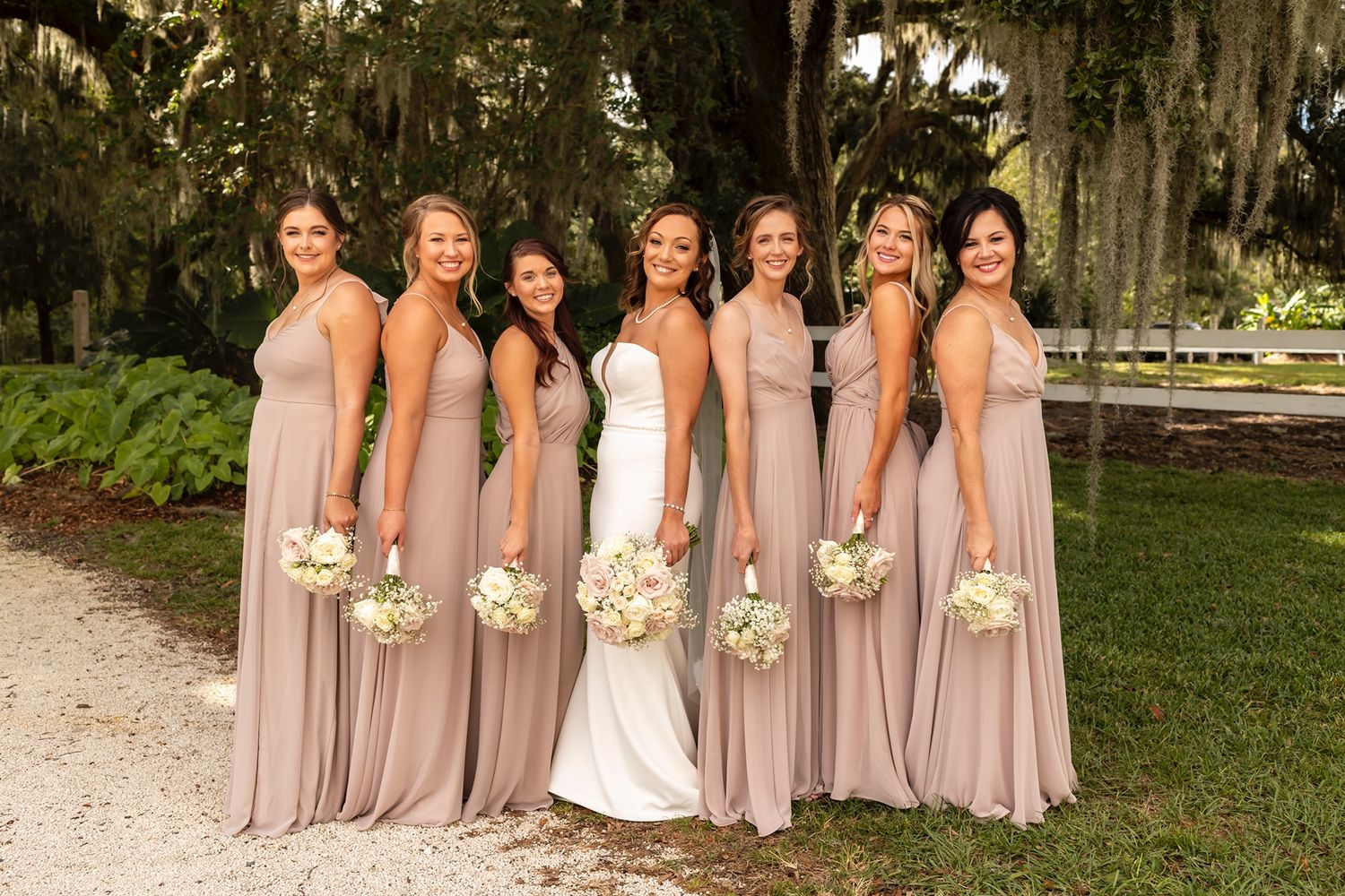 Bride in white wedding dress stands with six bridesmaids wearing flowing champagne colored gowns on a garden path.