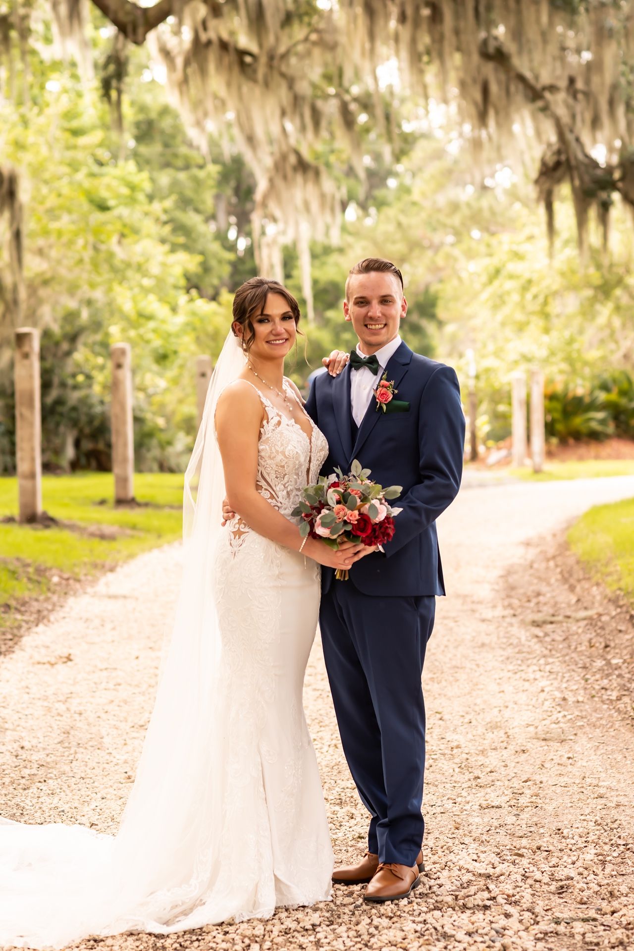 A couple in wedding attire stands together on a tree-lined path with Spanish moss overhead on their special day.
