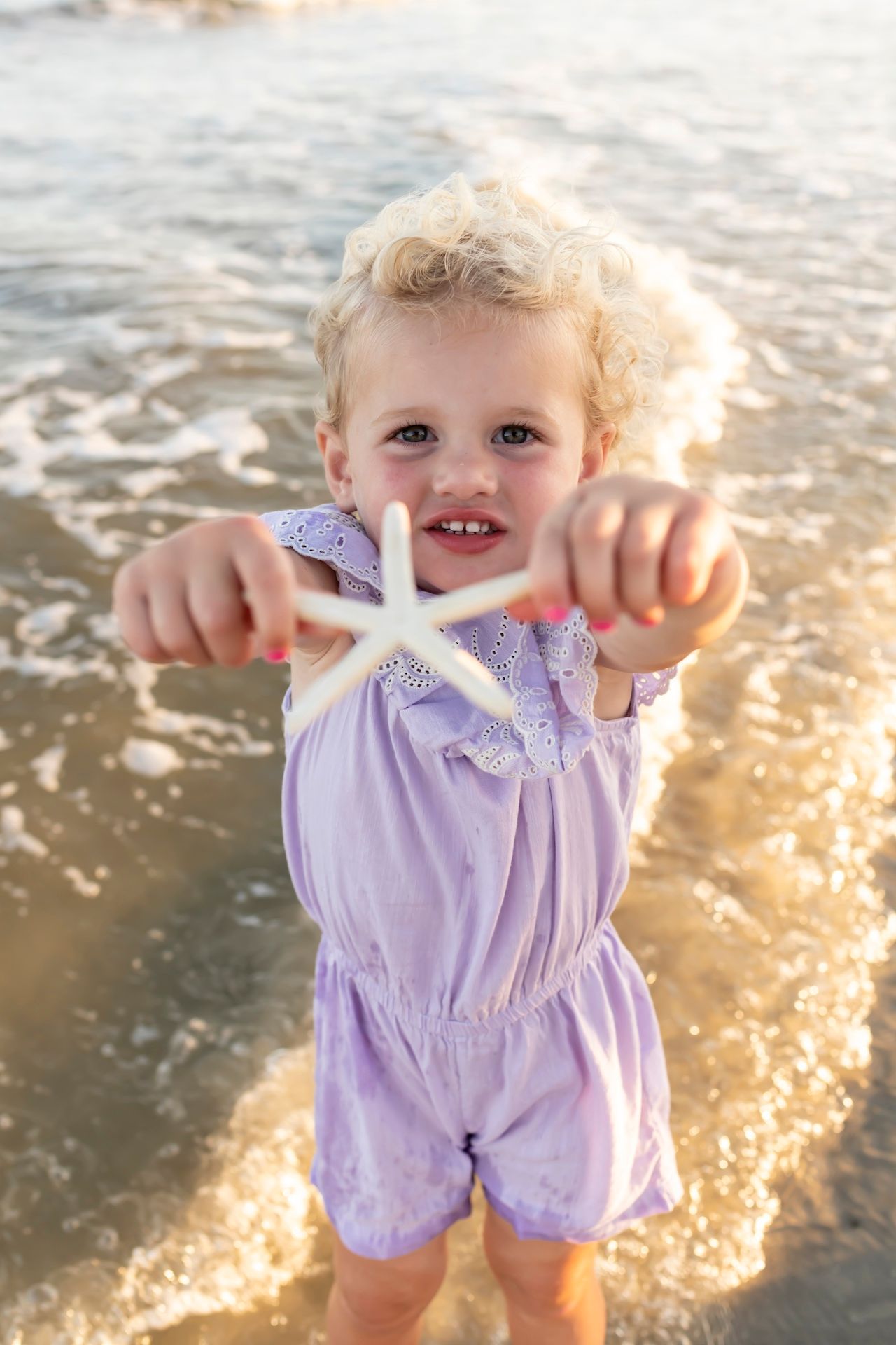 A small child in a purple outfit holds up a starfish on the beach at golden hour.