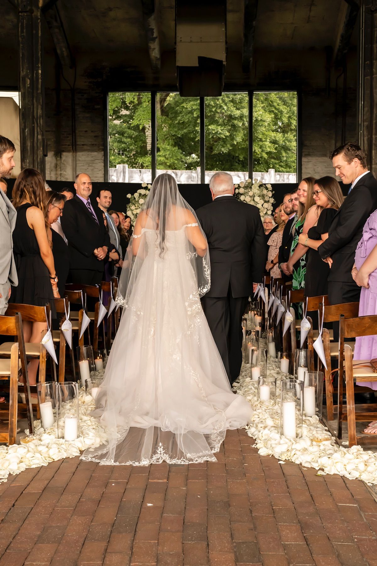 A bride in a flowing white dress and veil walks down a candlelit aisle at an indoor ceremony venue.