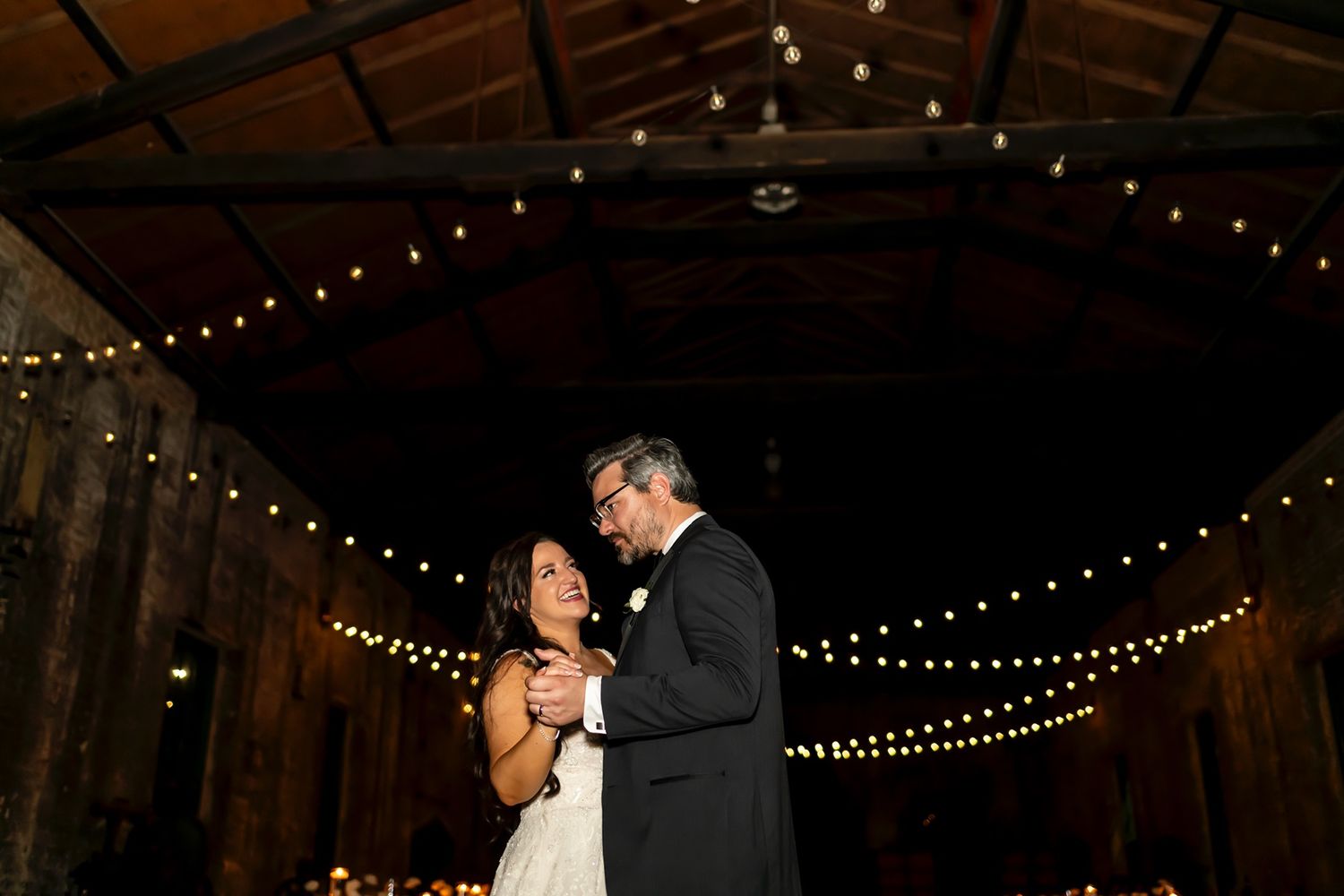 A couple shares an intimate dance under romantic string lights in a rustic barn venue during their wedding celebration.