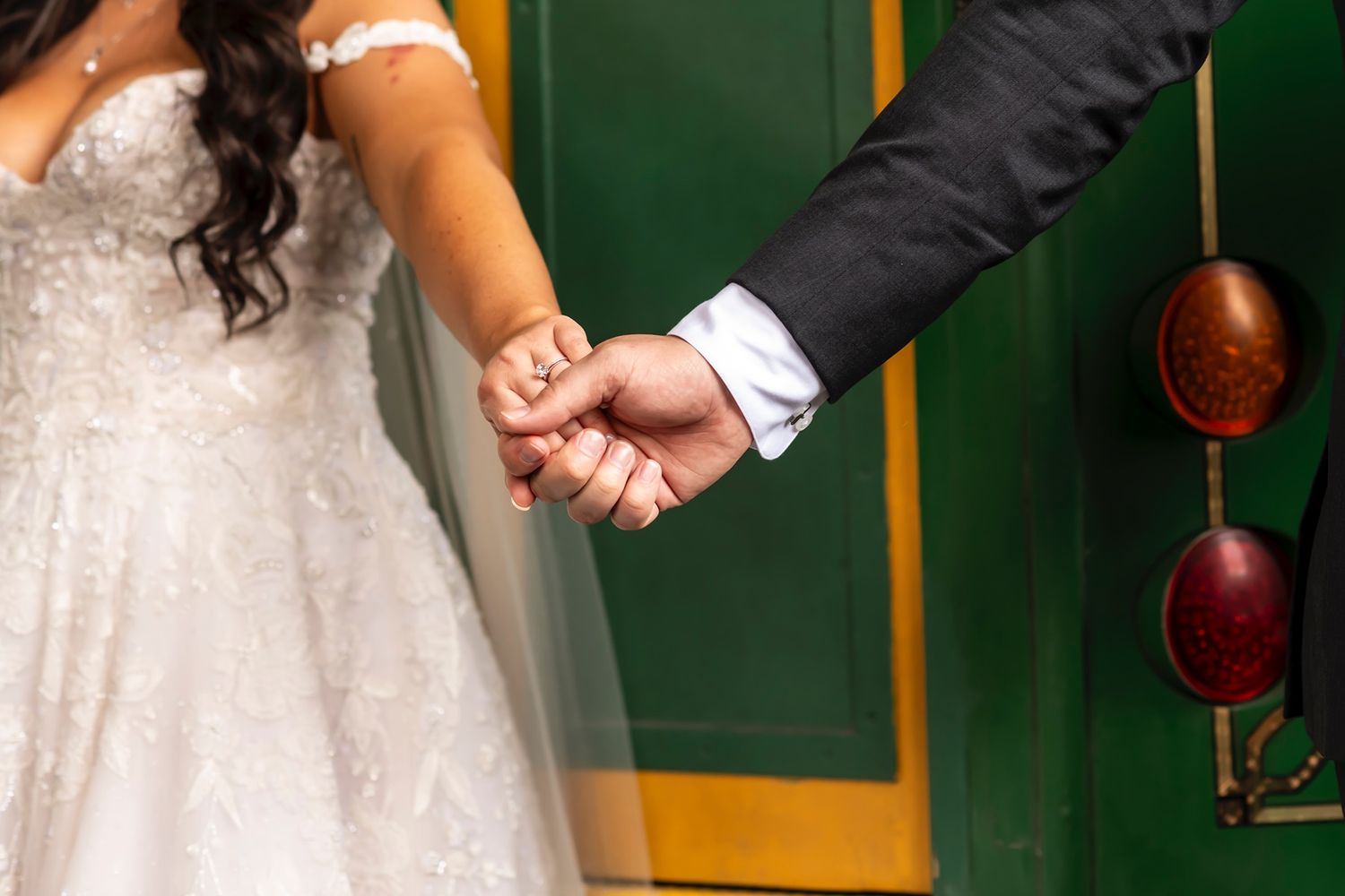 A close-up shot of newlyweds holding hands, showing a white wedding dress and formal suit sleeve against a green background.