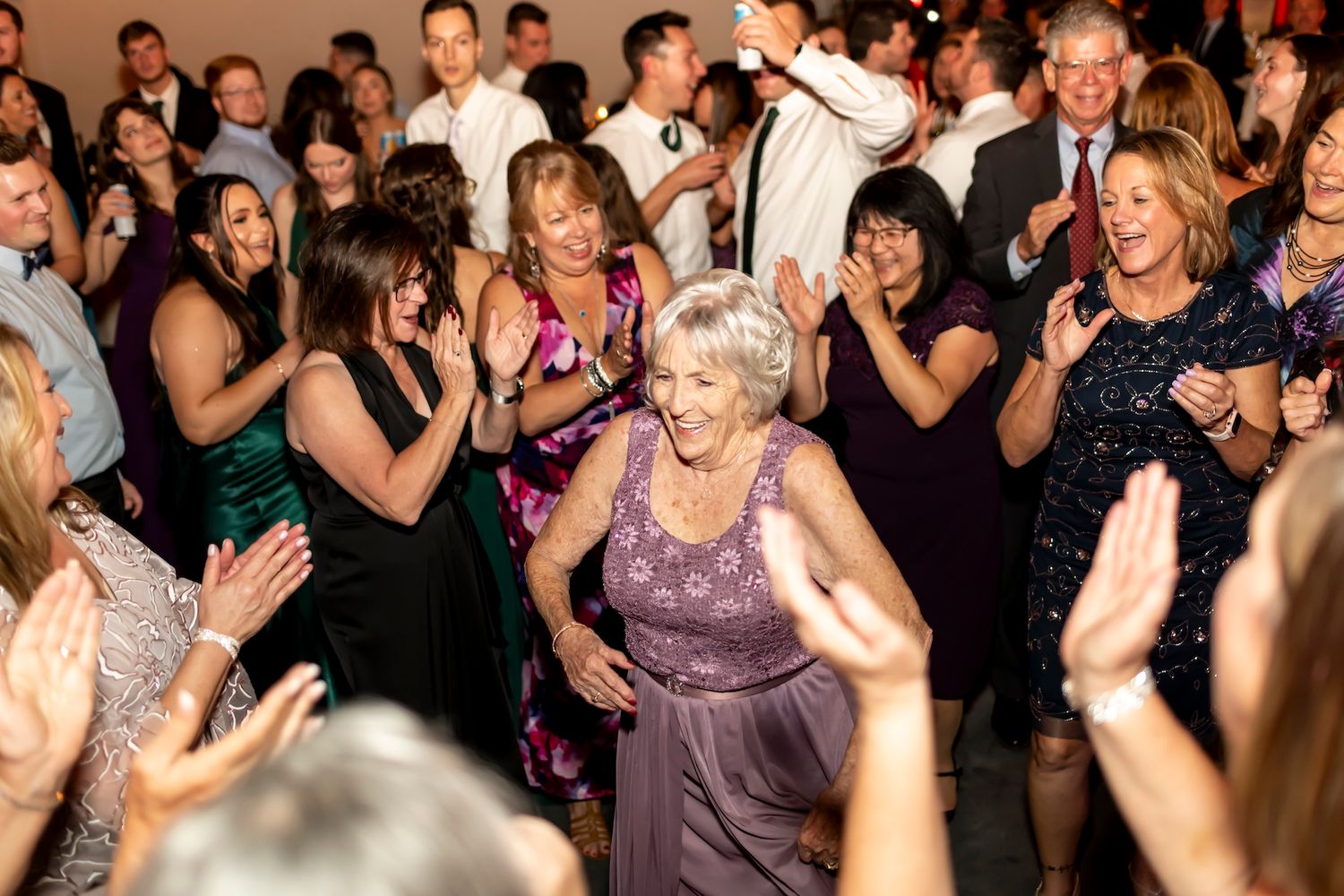 A group of party guests dance and cheer enthusiastically with someone in a purple dress at the center of the dance floor.
