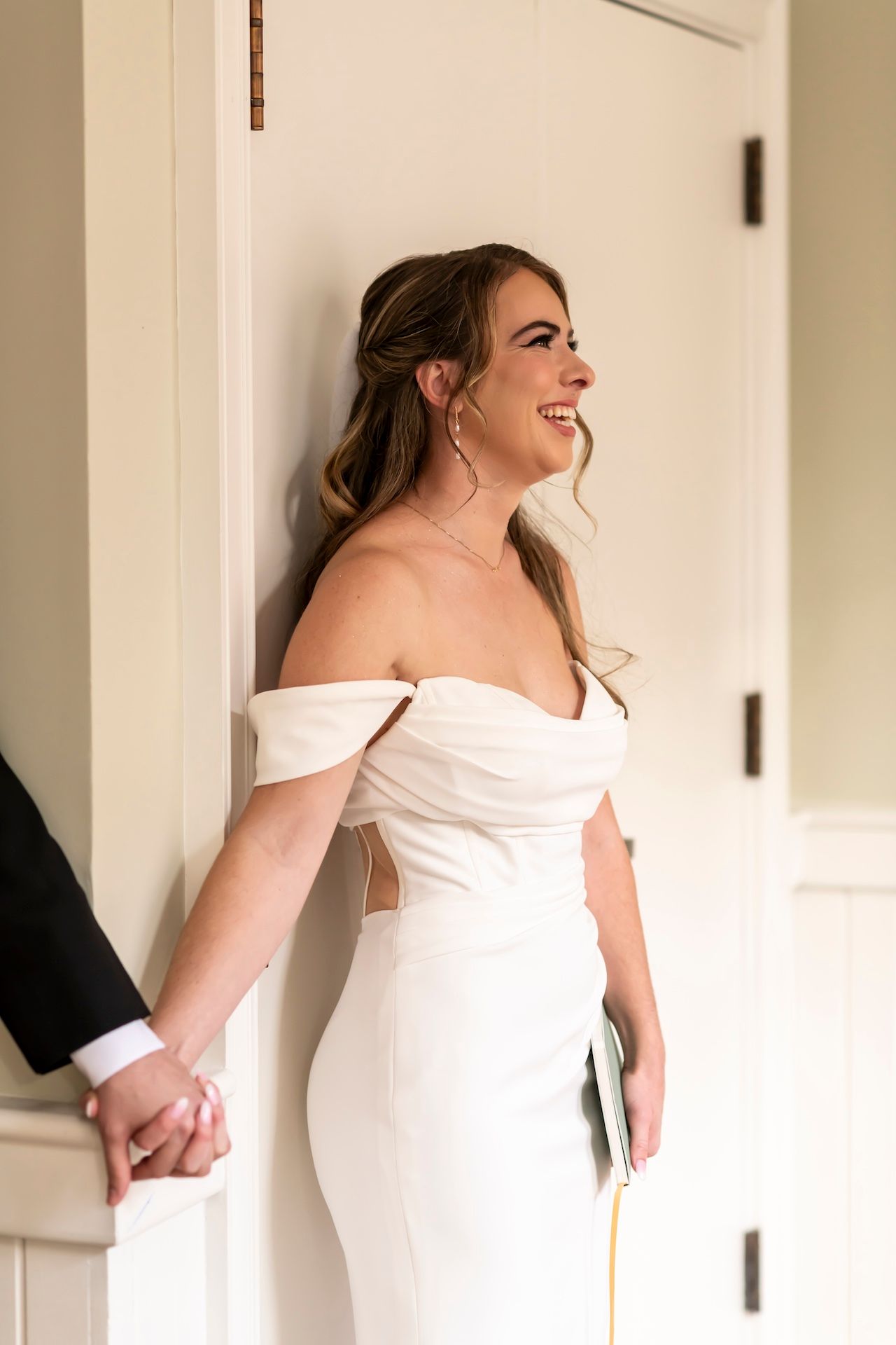 Joyful bride in off-shoulder white wedding dress laughs while holding hands with someone out of frame.