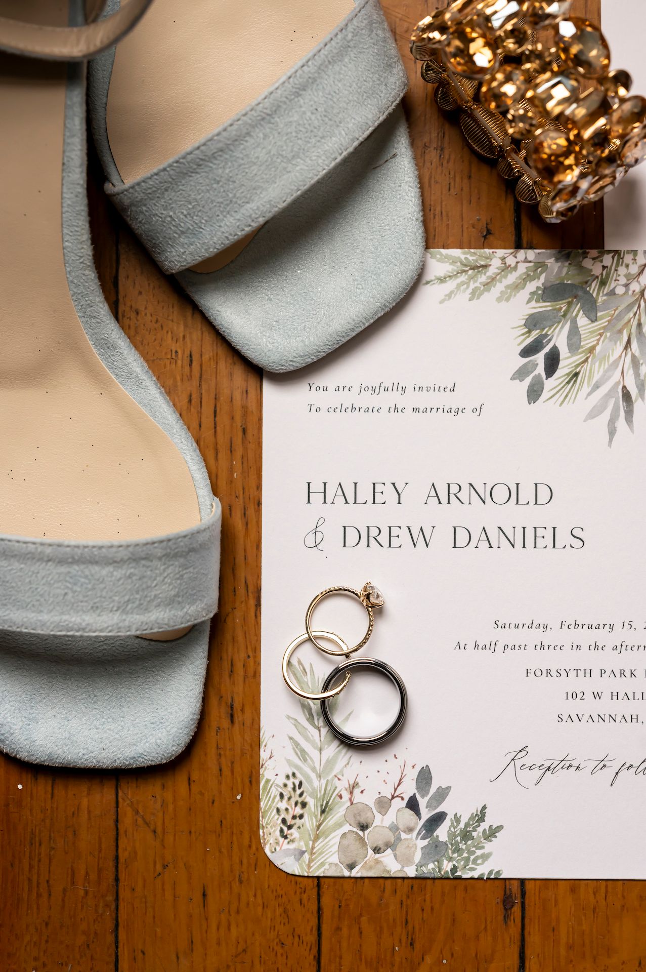Wedding invitation with floral design sits beside silver rings and blue heels on wooden table.