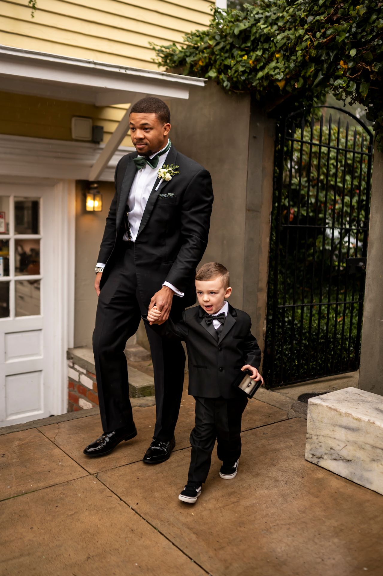 A well-dressed duo in matching black tuxedos and bow ties pose together outside a yellow house entrance.