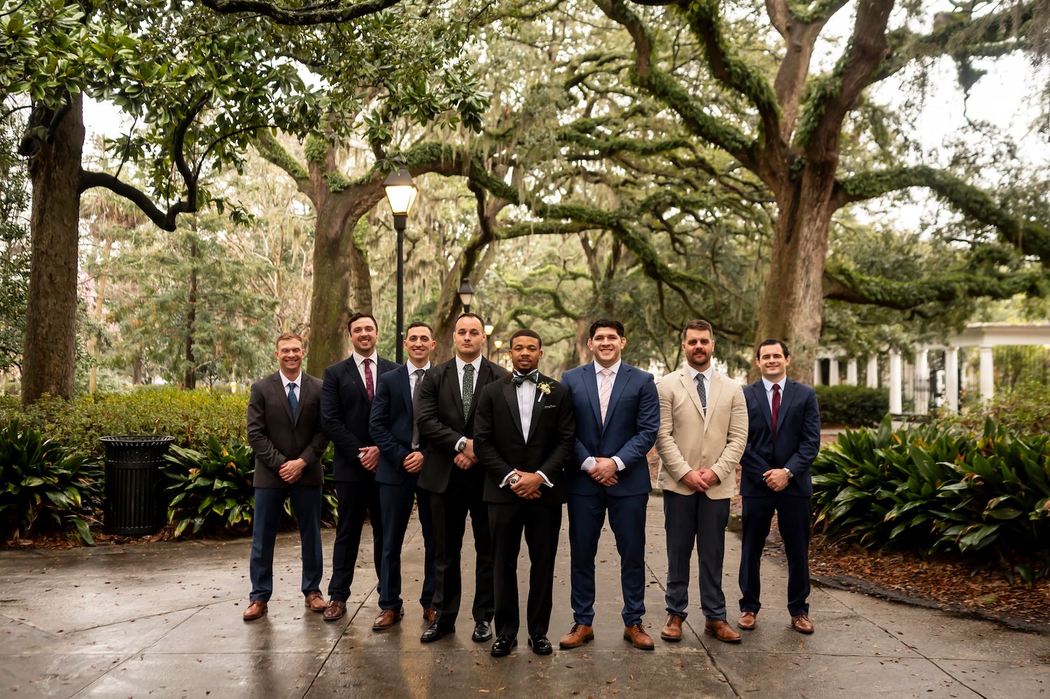 Wedding party group standing on path under majestic oak trees draped with Spanish moss in outdoor garden setting.