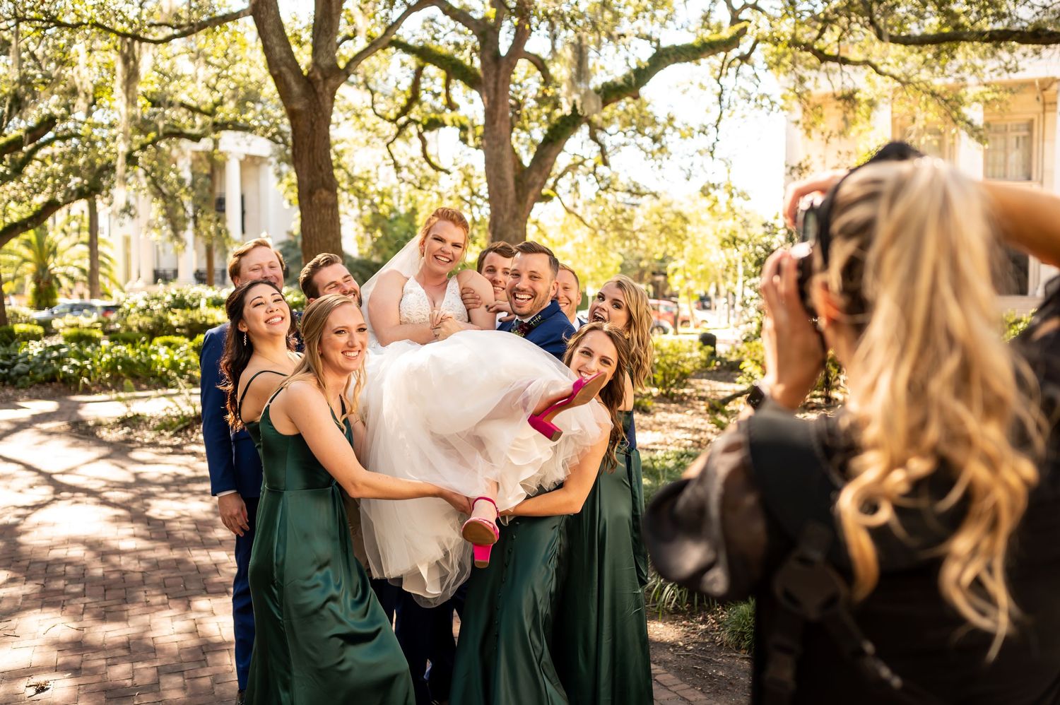 Photographer captures wedding party in emerald green dresses posing under moss-draped trees in a garden setting.