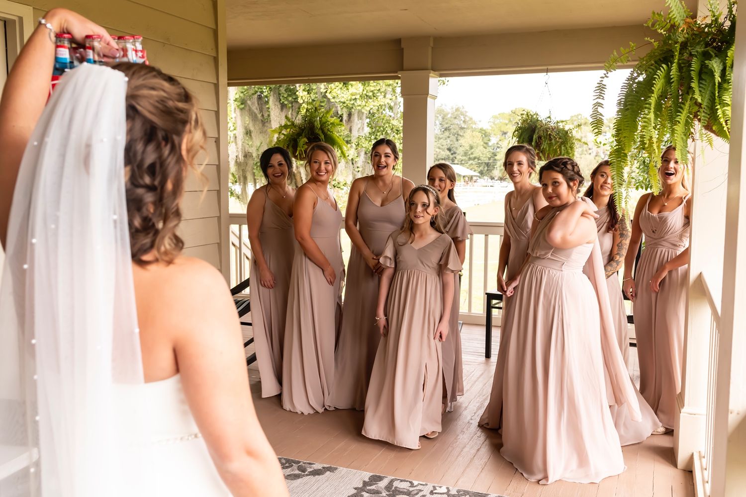 Bridesmaids in blush pink dresses gather on a porch to see bride in her wedding gown for the first look.