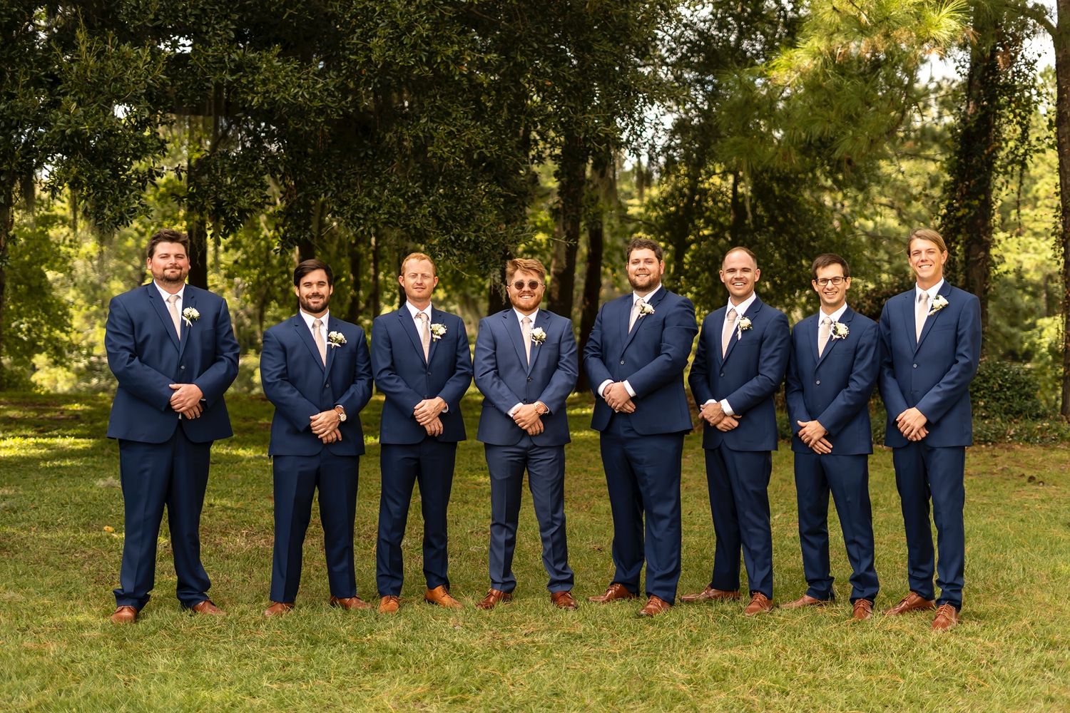 A group stands in a row outdoors wearing matching navy blue suits, white shirts and brown dress shoes at a formal event.