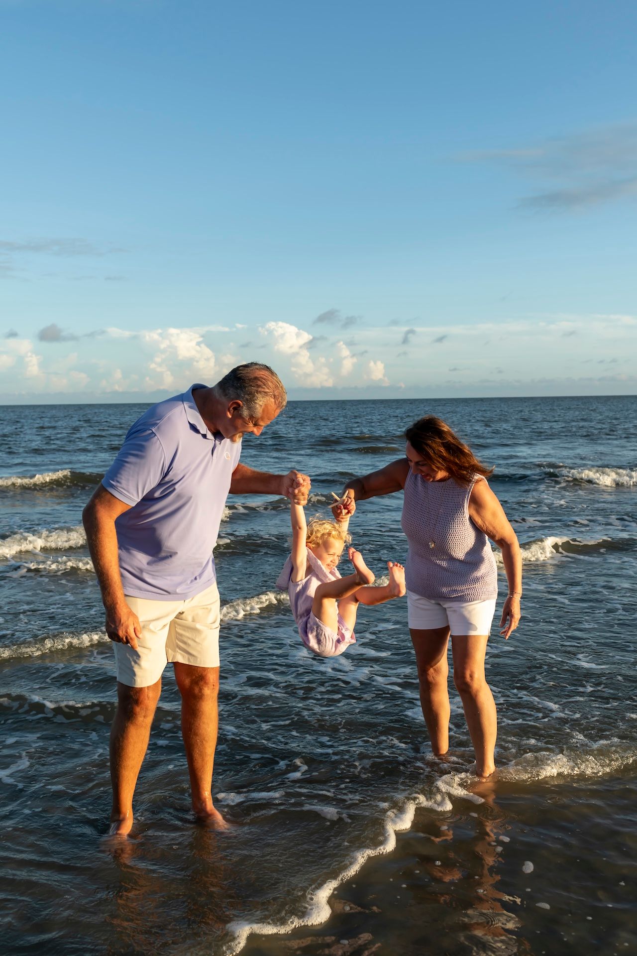 A couple swings a small child while enjoying a sunset walk along the water's edge at the beach.