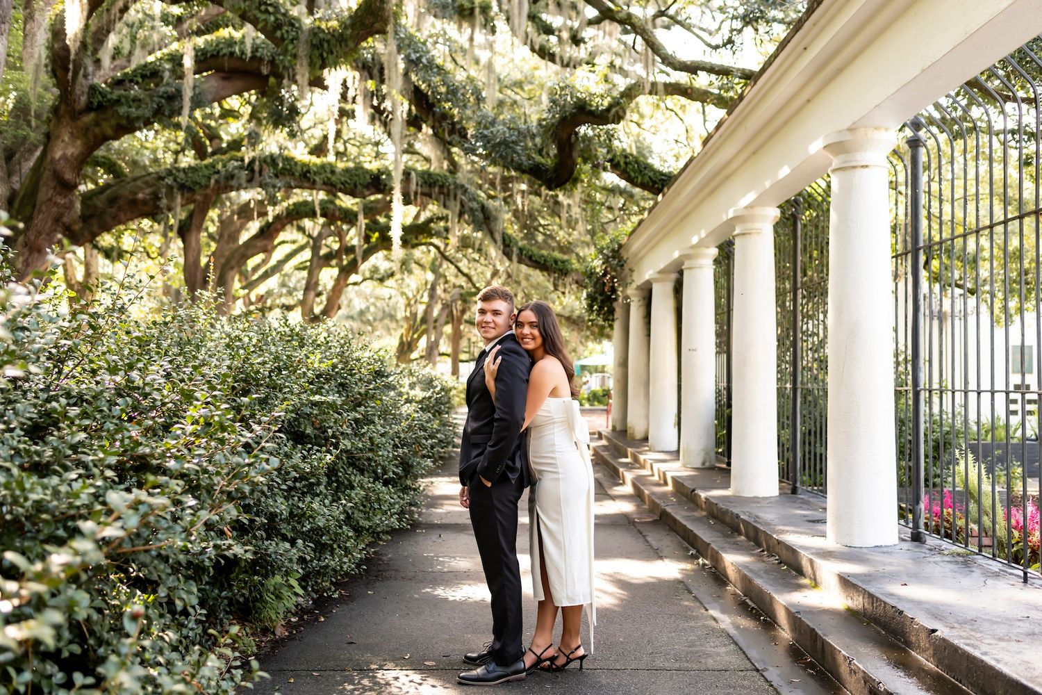 A couple stands in an elegant white columned portico walkway beneath moss-draped oak trees.
