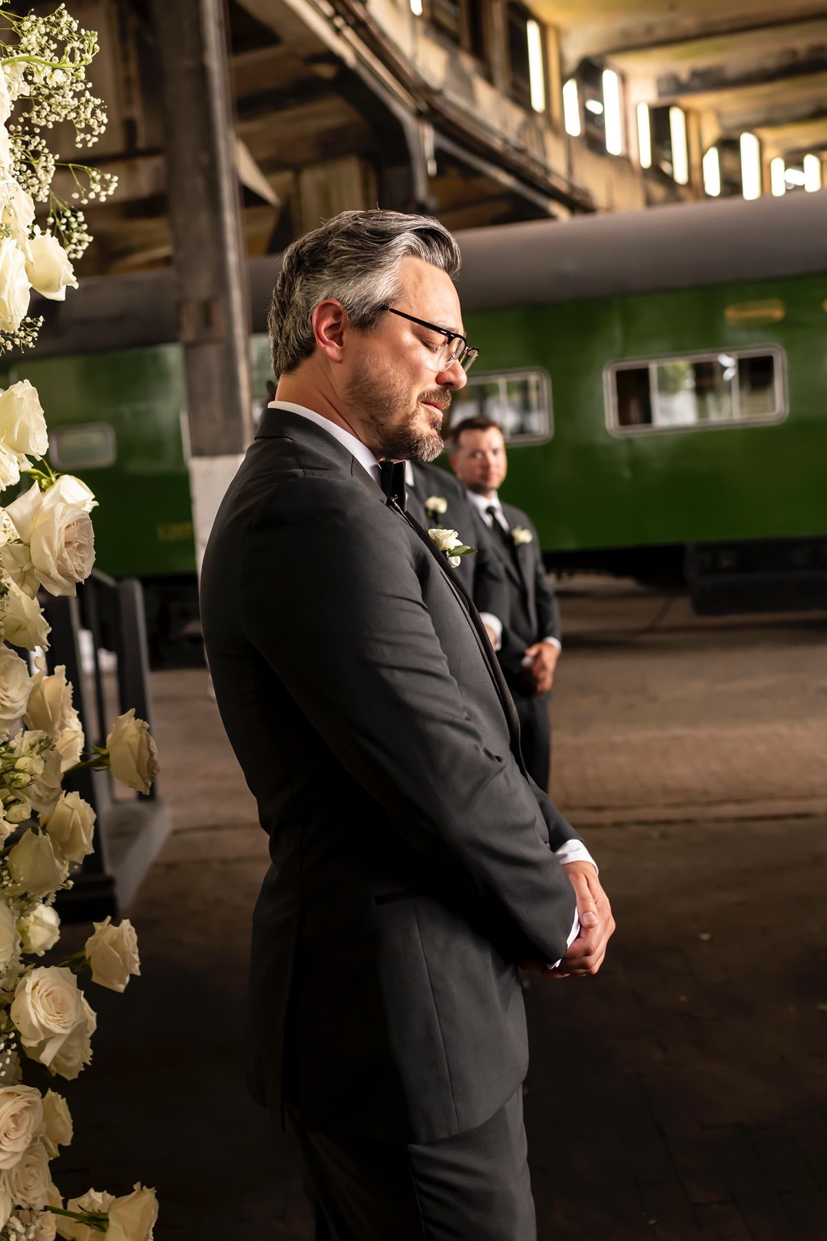 A groomsman in a black suit and glasses stands beside white floral arrangements inside a rustic venue.