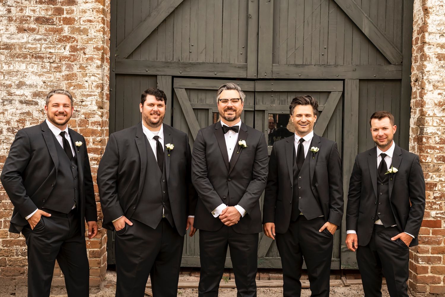 Groomsmen in classic black suits pose together smiling in front of rustic brick wall and wooden barn doors.