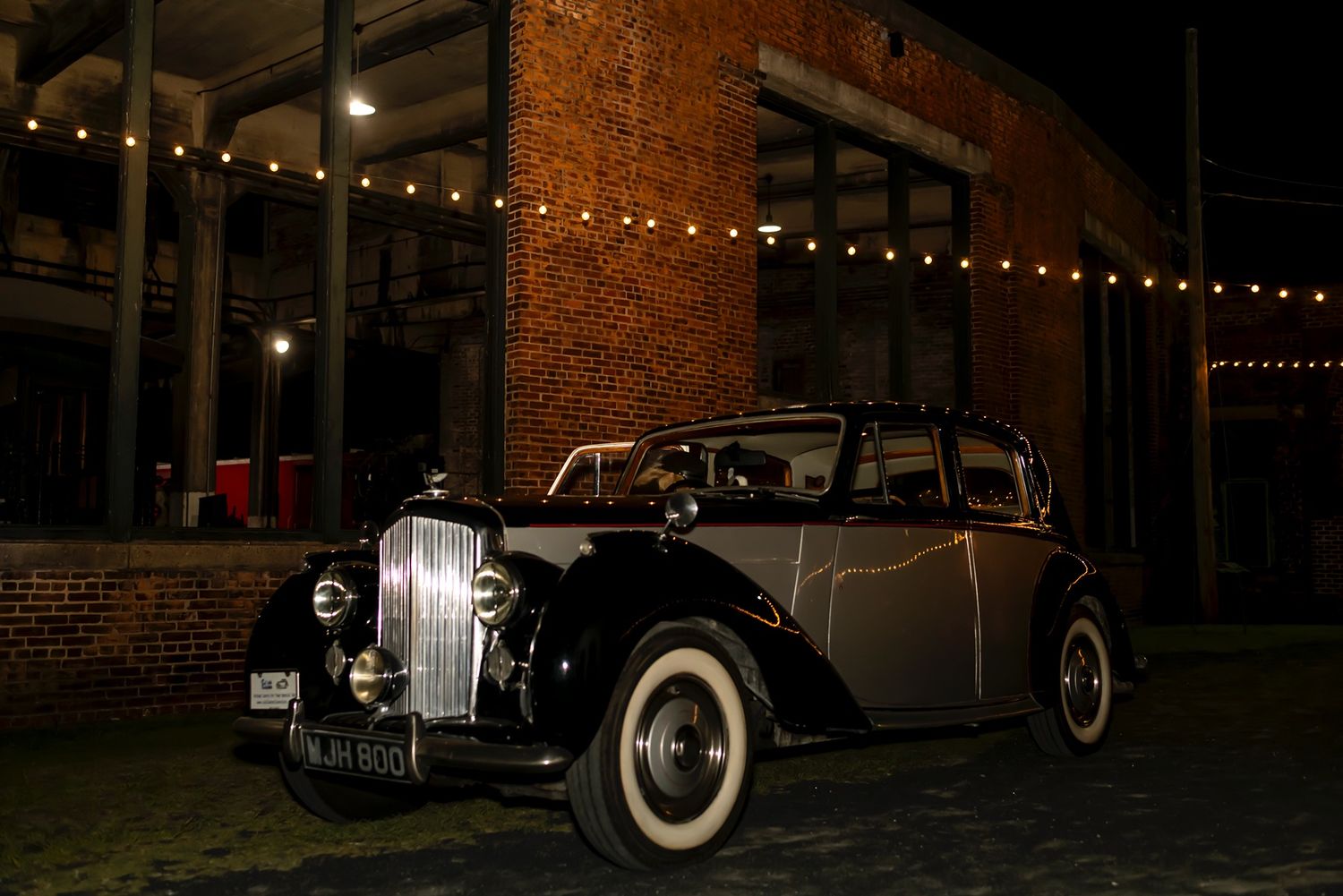 A dark classic vintage car parked in front of a brick building illuminated by warm string lights at night.