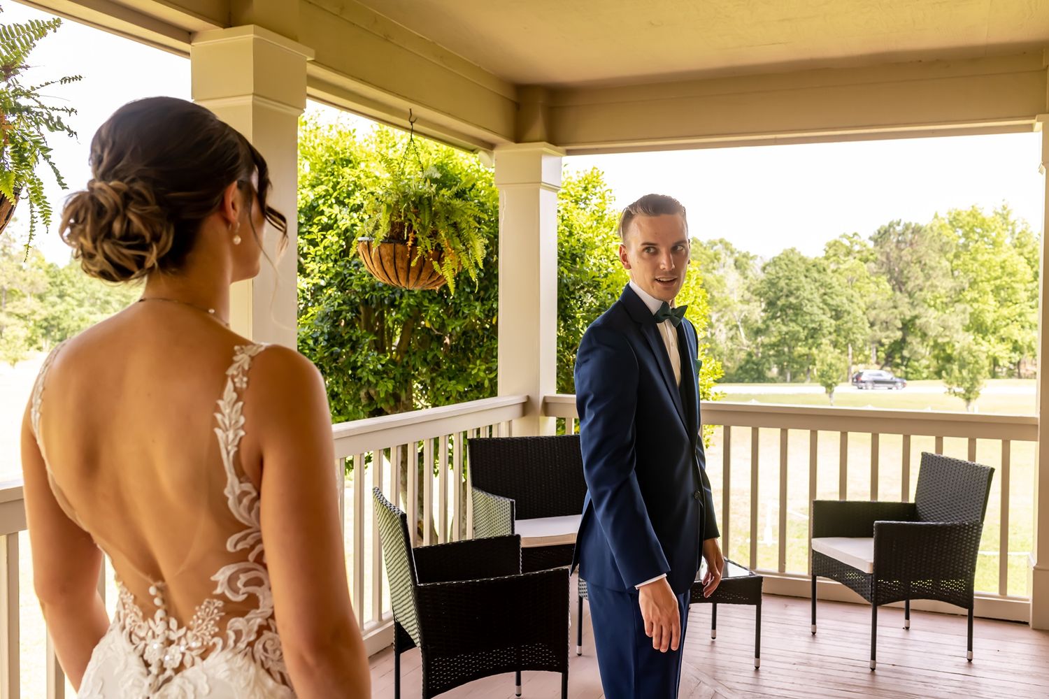 First look between a bride in a lace back dress and a groom in a navy suit on a sunny outdoor porch.