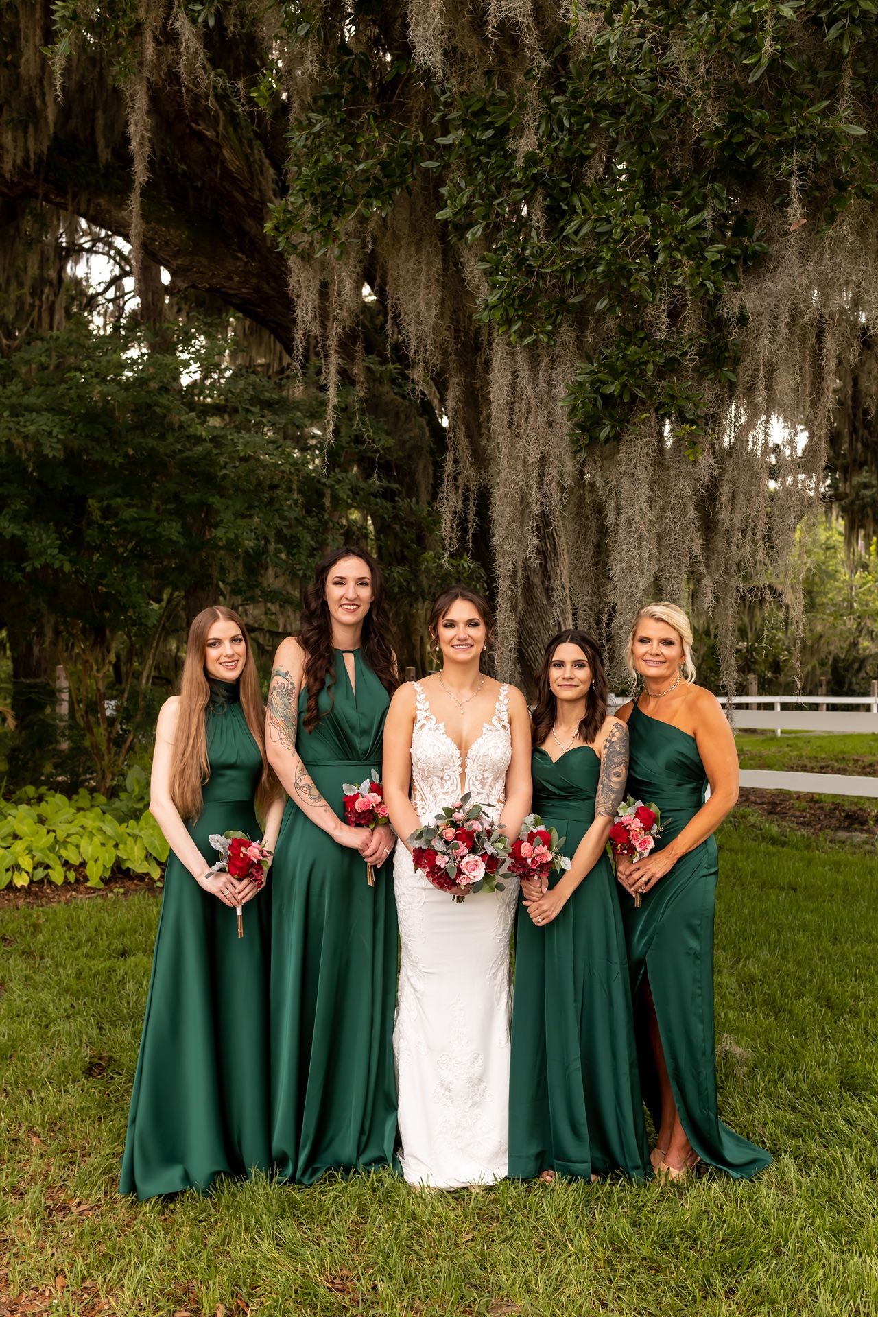 Wedding party poses outdoors with bridesmaids in emerald green dresses and bride in white gown under Spanish moss trees.